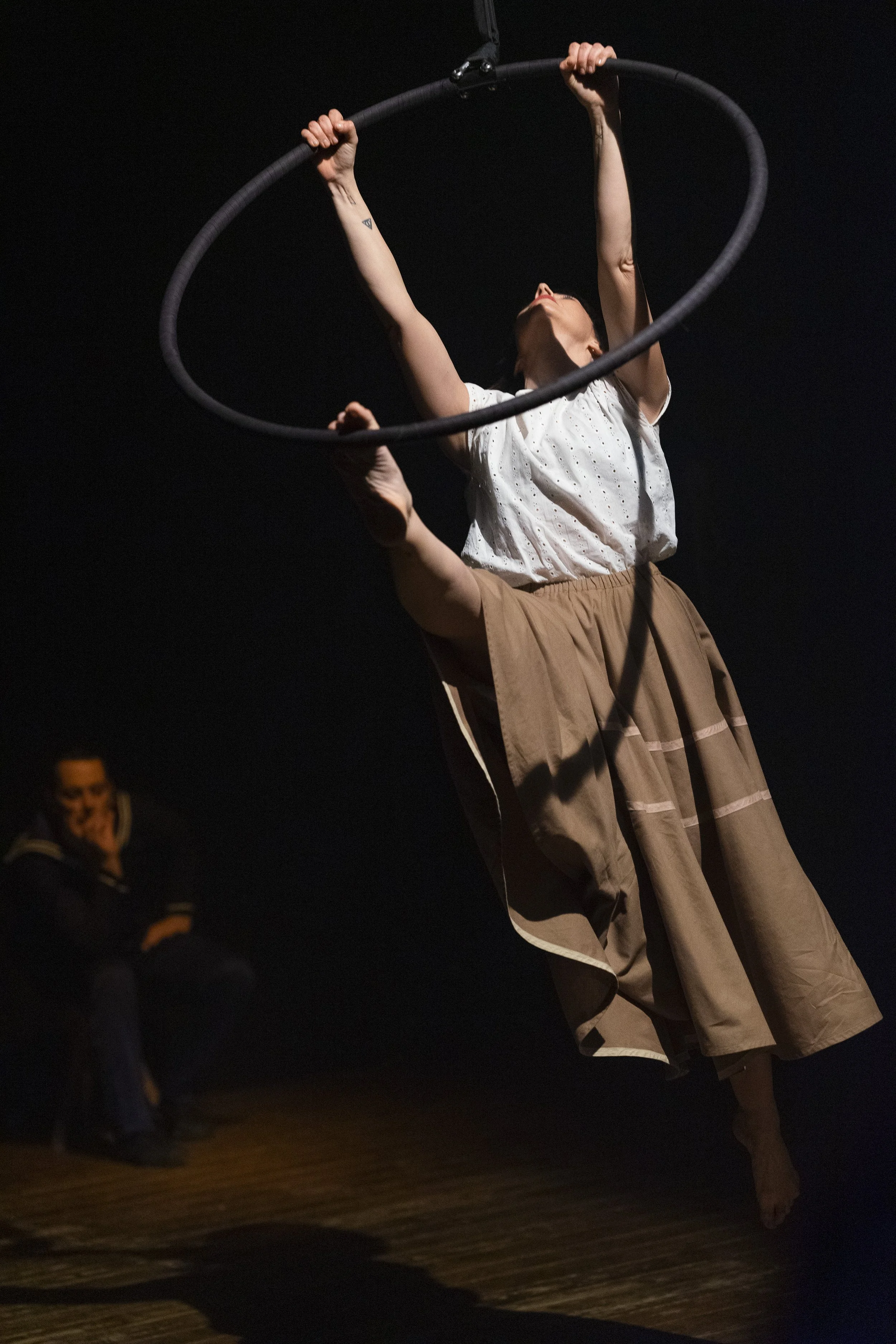 Una donna che esegue un'performance con un hula hoop in un teatro oscuro, indossa un abito largo marrone e una camicia bianca, mentre un uomo seduto sul lato sorride guardando.