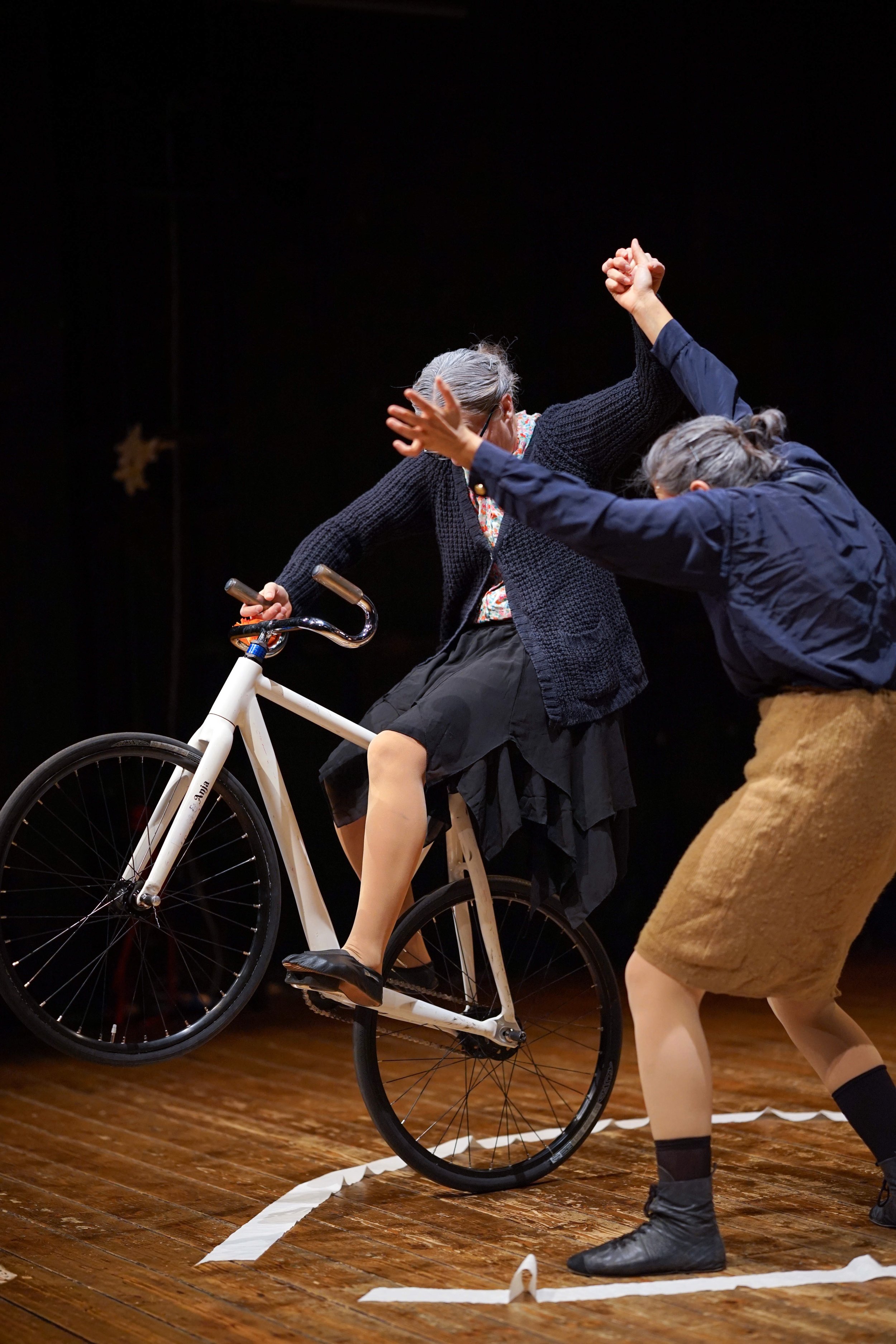 Due persone stanno esibendo uno spettacolo di teatro o danza, con una di loro seduta su una bicicletta e l'altra che la sostiene, su un palco di legno.