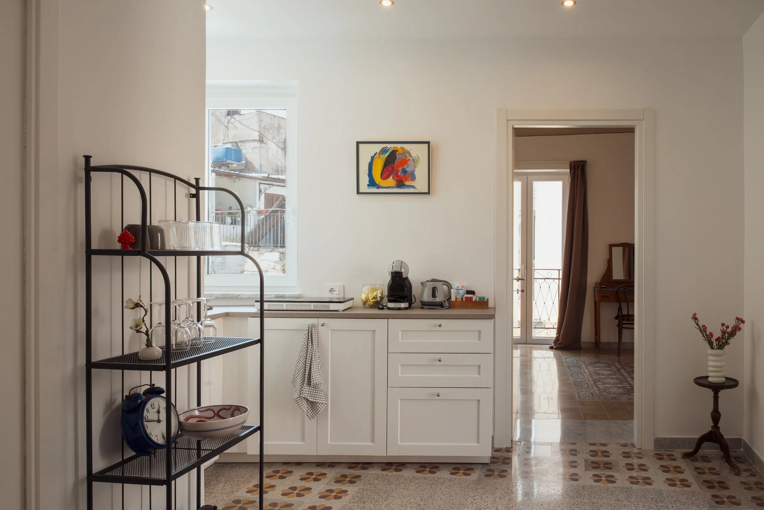 Interno di una cucina con mobile bianco, ripiano con macchina da caffè, tostapane, e vari oggetti. Sopra il mobile c'è un quadro astratto, a sinistra una finestra e a destra un passaggio verso un'altra stanza con balcone, tavolo e sedia. Sul paviment