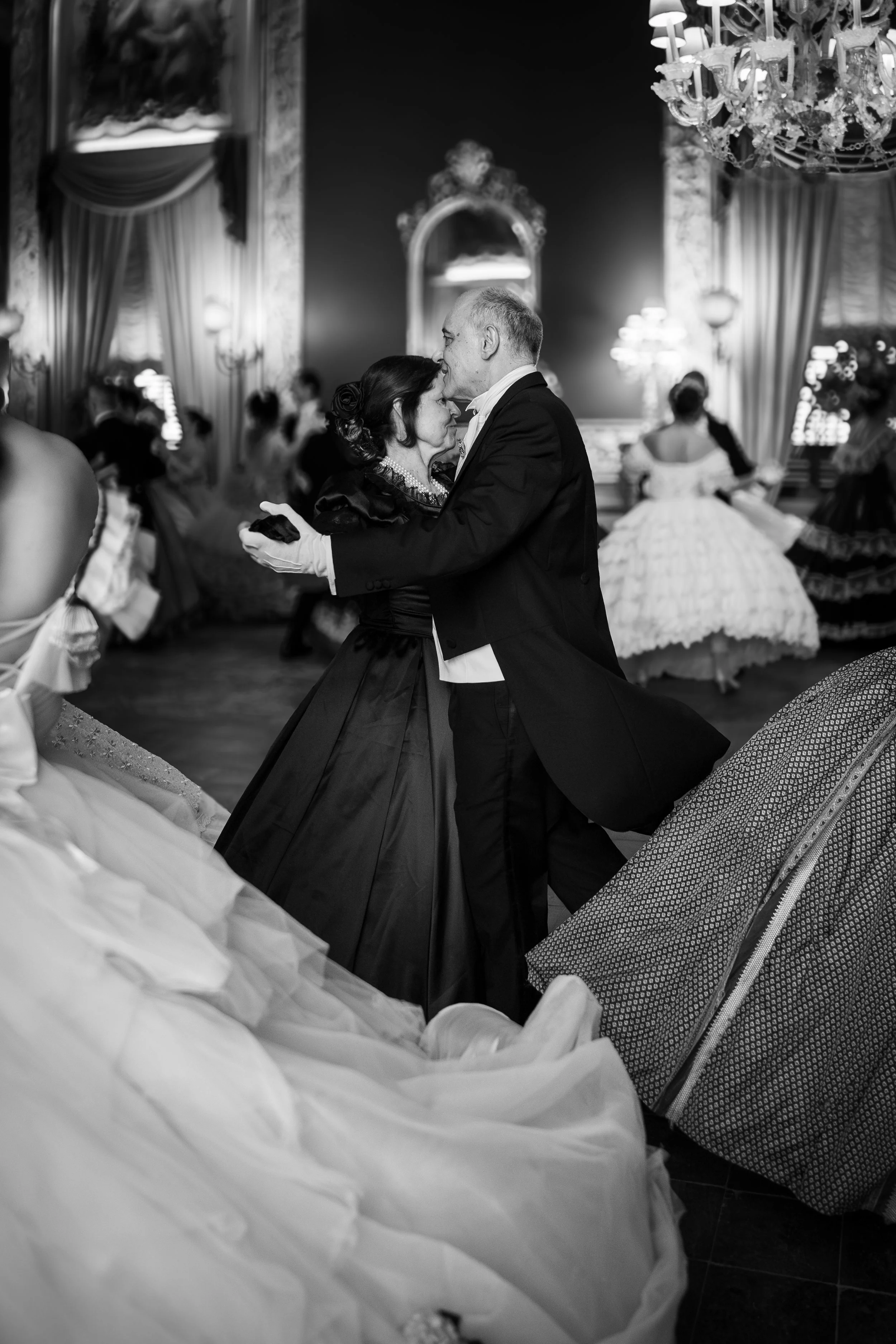 Una coppia in abiti eleganti danza nel centro di una sala ornata con luci soffuse, lampadari e grandi tende, con altre persone vestite in abiti storici che fanno parte di una scena teatrale o di un evento storico.