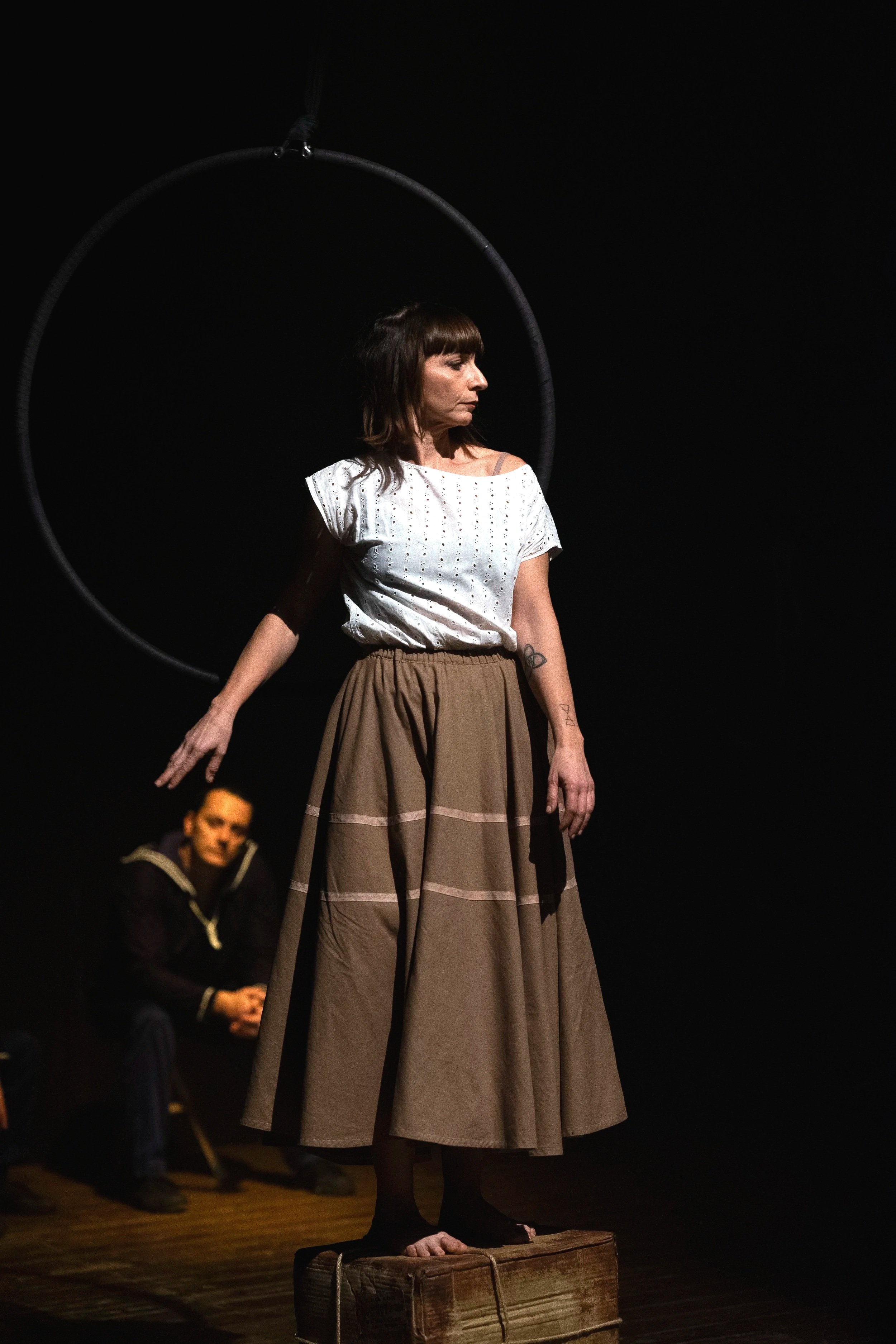 Una donna sta in piedi su una scatola, con un cerchio sospeso sopra di lei, mentre un uomo seduto in background la osserva. La scena sembra essere parte di una performance teatrale.