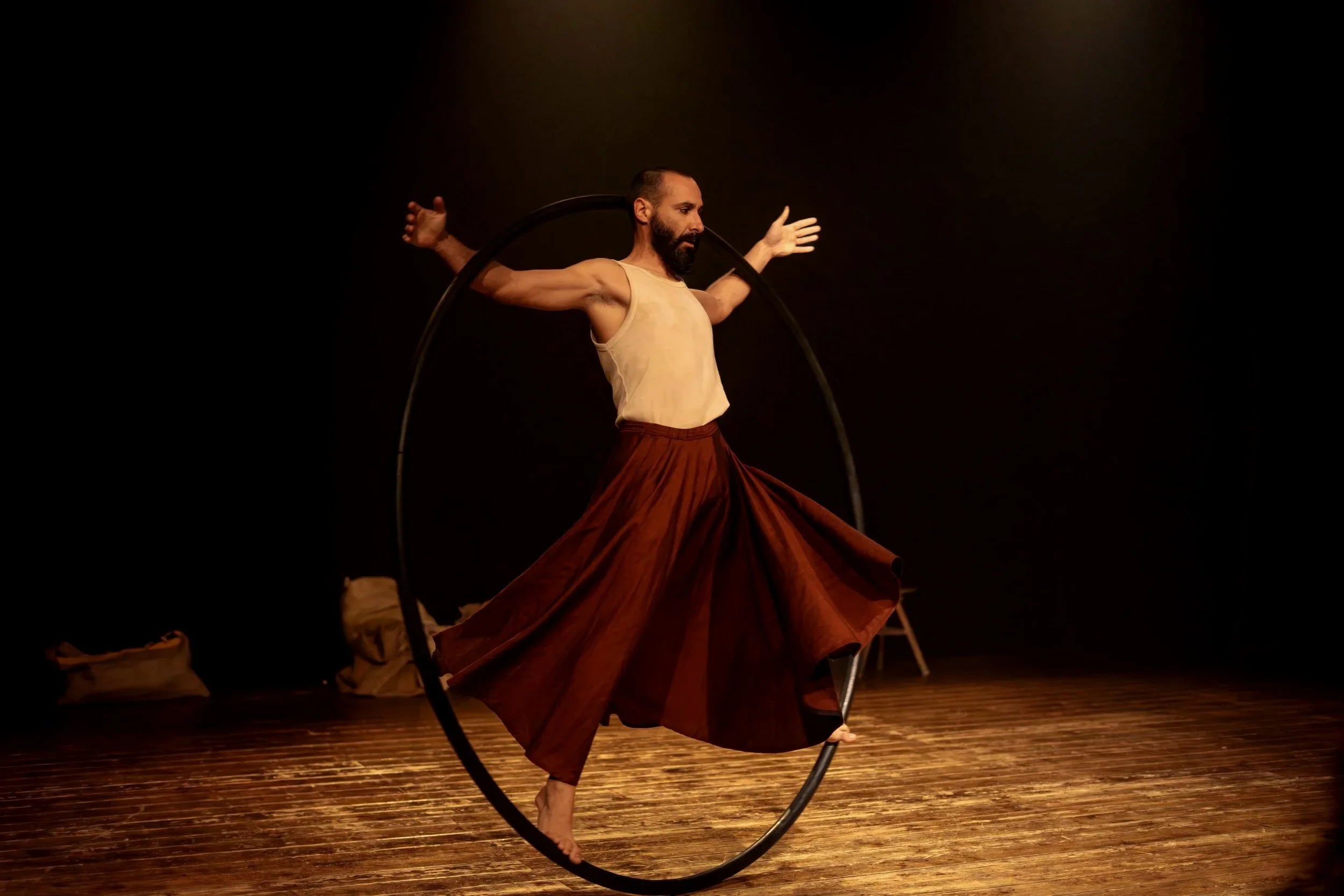 Uomo che esegue acrobazie con un cerchio, vestito con pantaloni ampi e senza maglietta, sul palco in un teatro