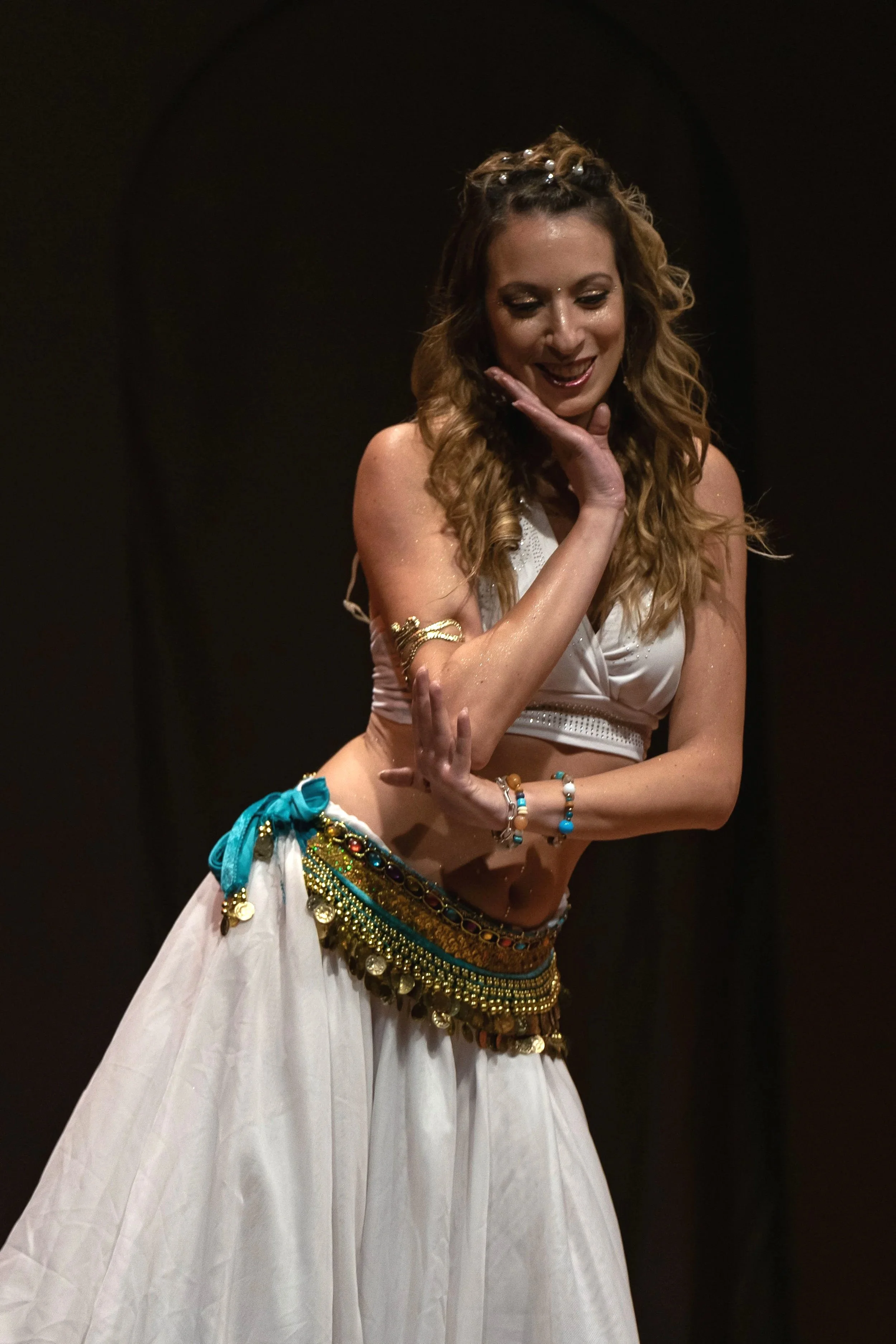 Una donna con capelli riccioli castani, indossa un abito bianco e accessori colorati, e fa una danza.