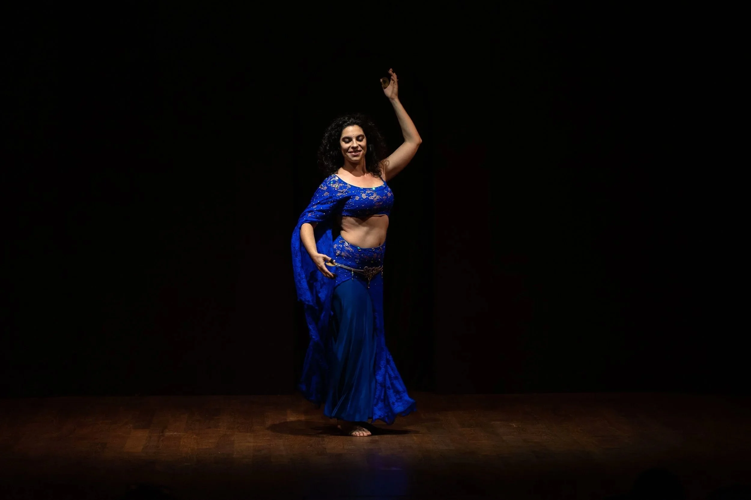 Donna che balla un ballo di danza del ventre su un palco con sfondo nero, indossa un vestito blu con dettagli di pizzo e gioielli.