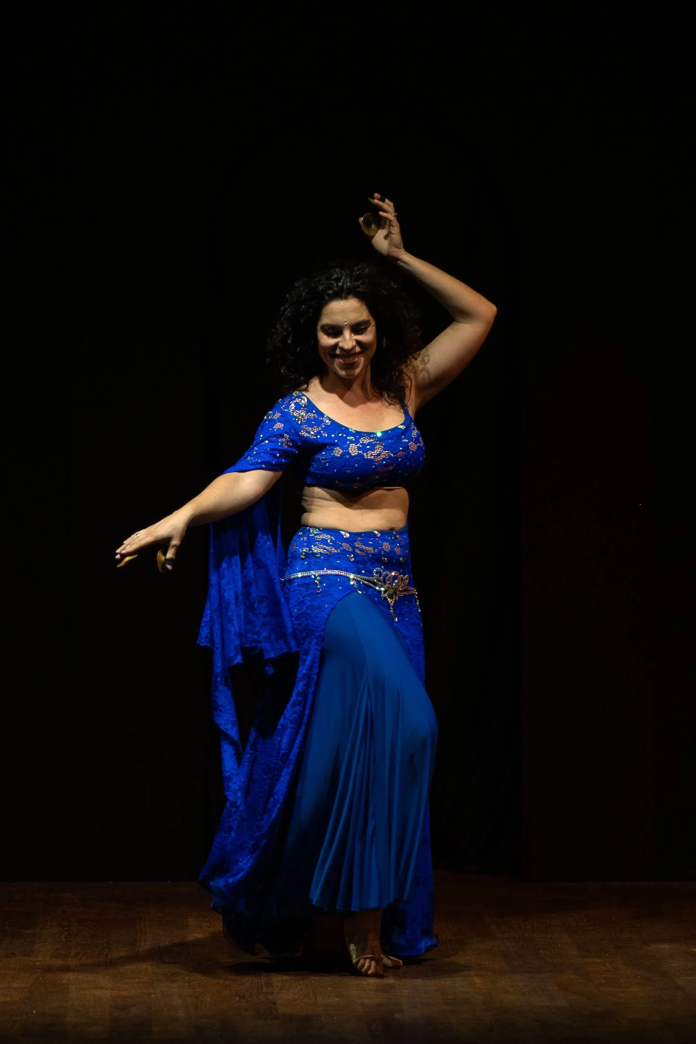 Donna che danza in un costume blu con dettagli in oro, sul palco con sfondo nero, che sorride