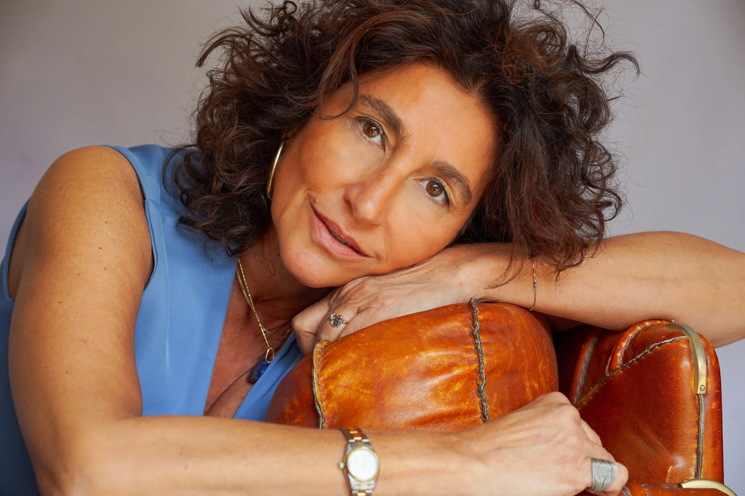 Donna con capelli ricci bianchi appoggia la fronte su un banco di legno, indossa un top blu e gioielli d'oro, guarda in camera con un sorriso e un'aria rilassata.