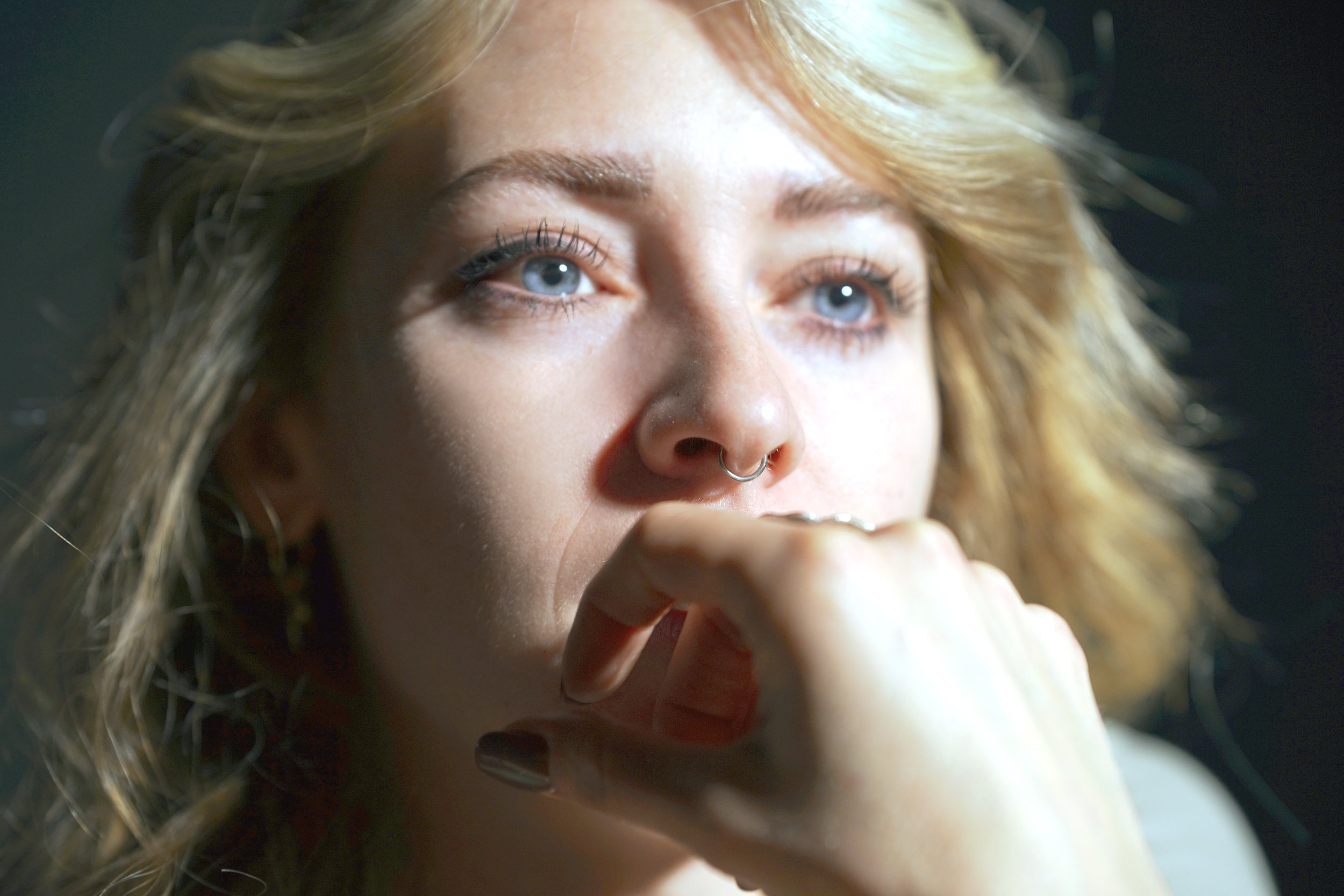 Primissima piano di una donna con capelli ricci biondi, occhi azzurri e septum piercing, che si poggia il mento sulla mano, in un'immagine ravvicinata.