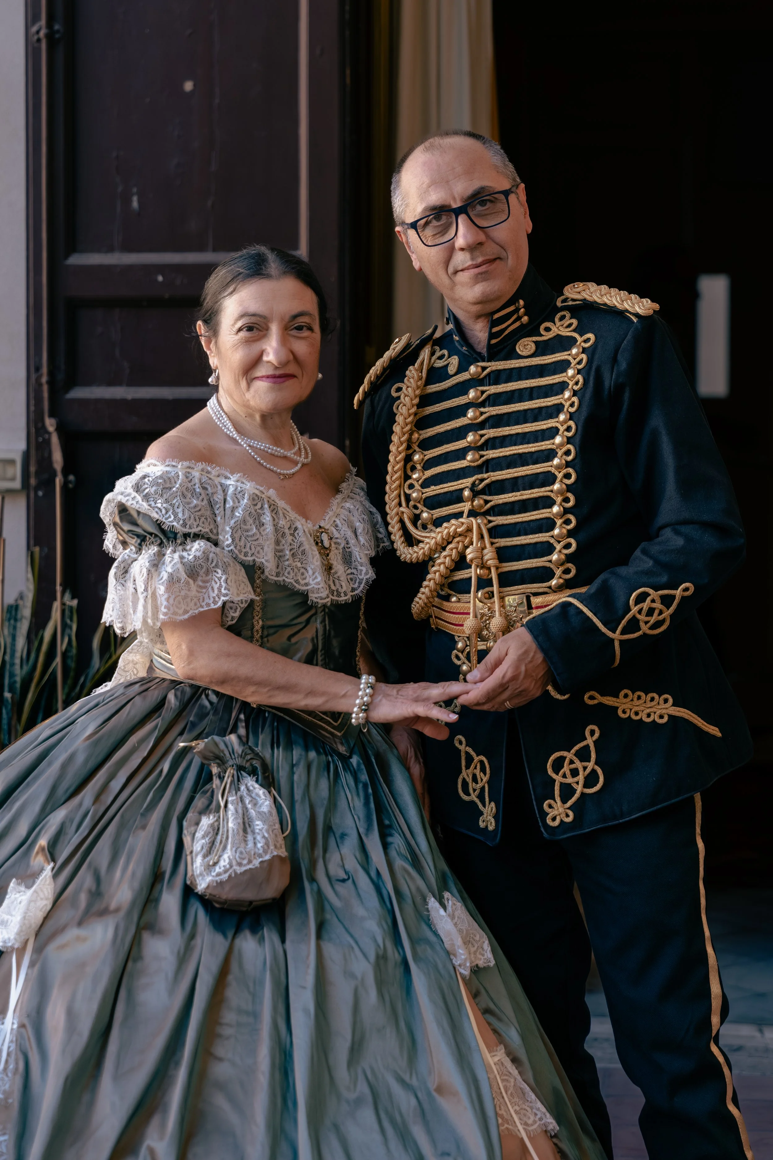 Una coppia di persone in abiti storici, una donna con un abito lungo con balze di pizzo e perle e un uomo con un'uniforme militare decorata con fili d'oro e bottoni.