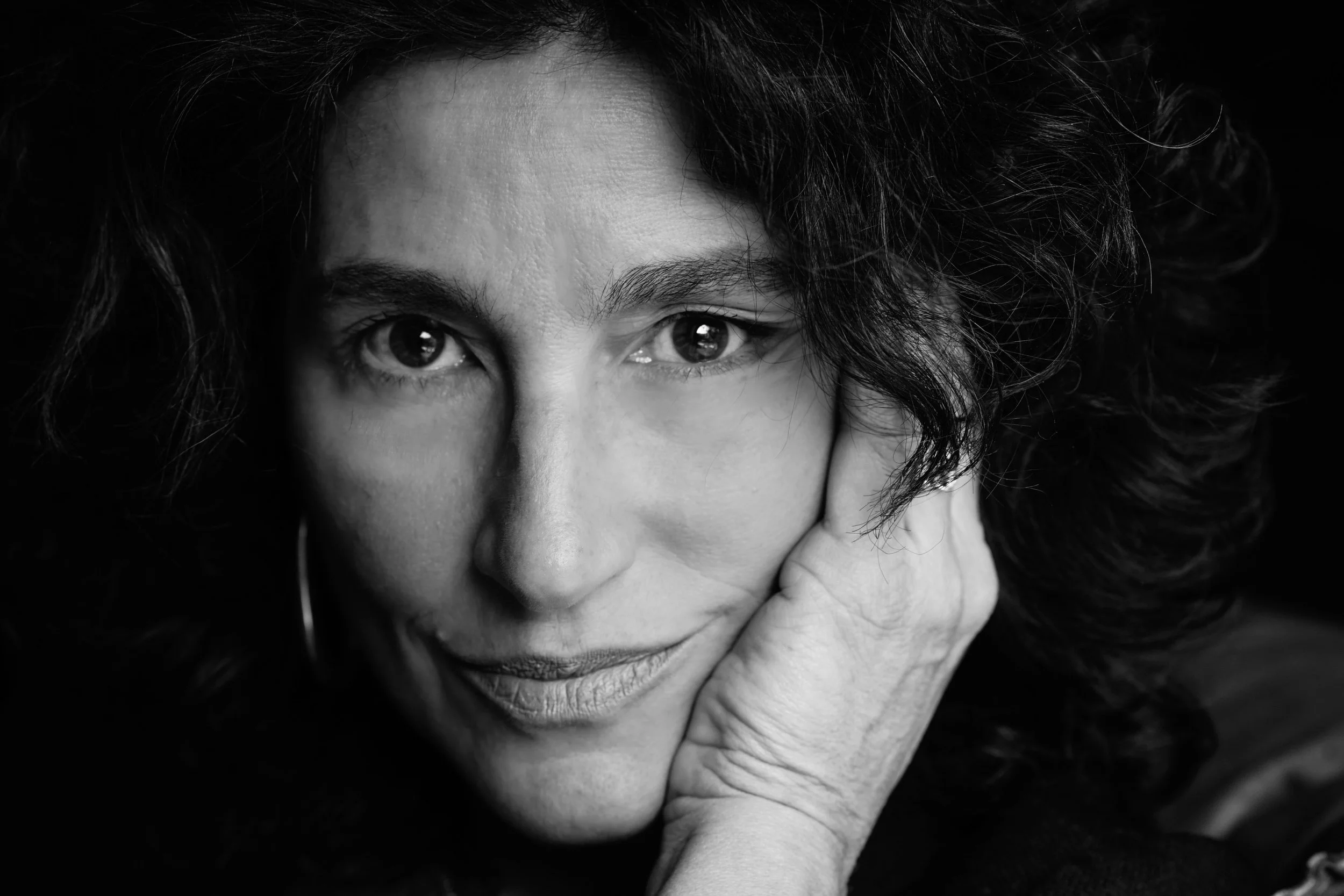 Ritratto in bianco e nero di una donna con capelli ricci, che sorride leggermente, appoggiando il viso sulla mano.