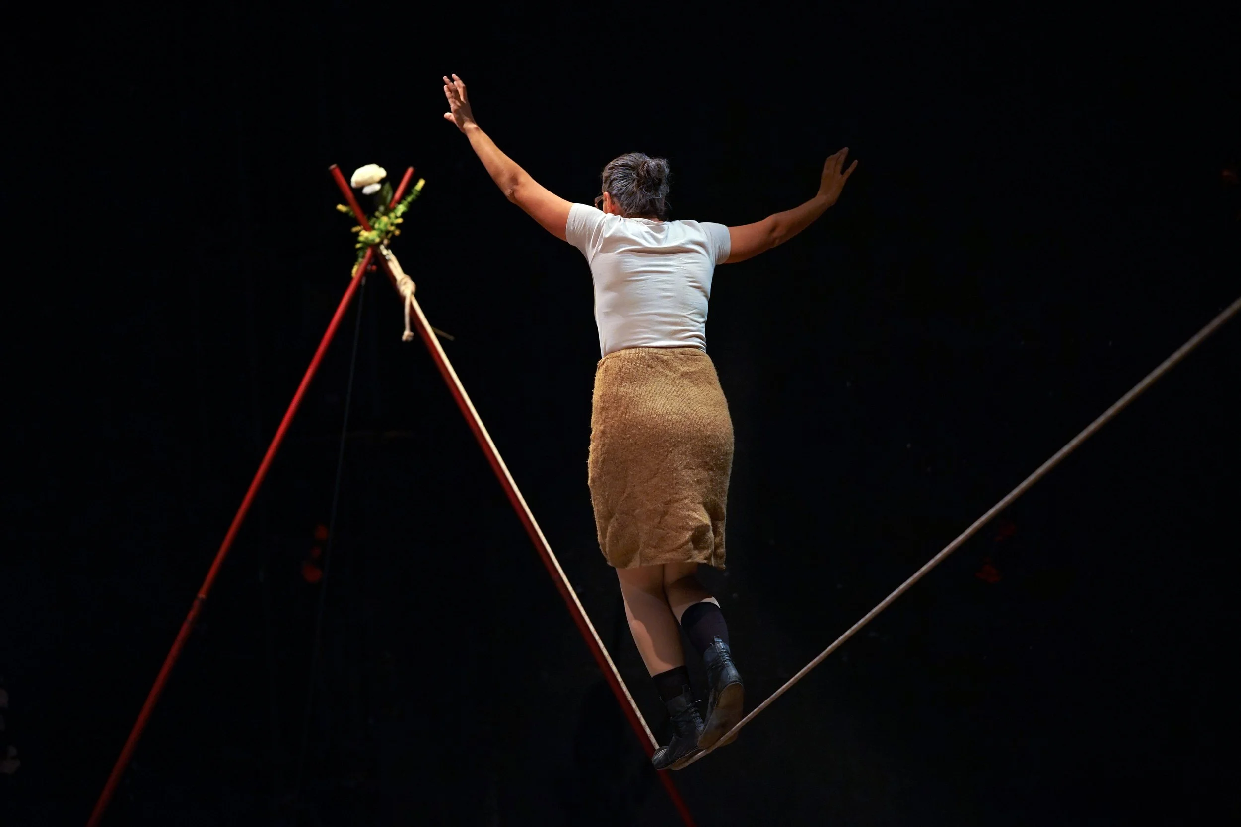 Una donna esegue un numero di equilibrio su una corda sospesa in aria, con le braccia aperte e un supporto decorato con fiori e bastoni di fronte a lei, sullo sfondo nero.