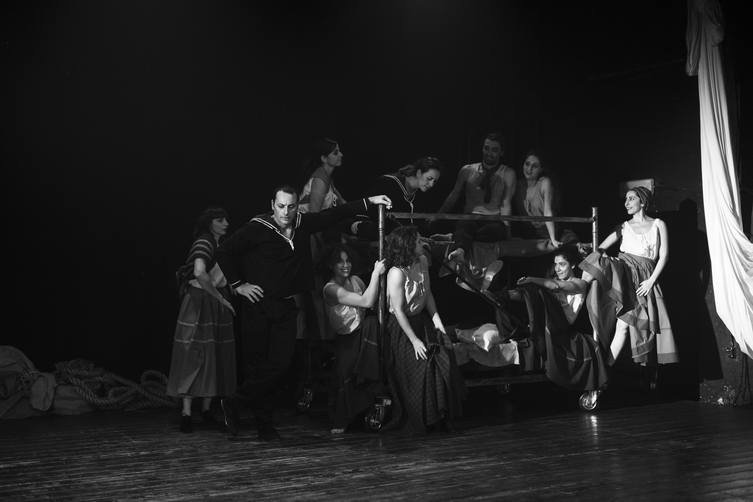 Foto di una scena teatrale in bianco e nero con diversi attori in costumi vintage, alcuni seduti e altri in piedi, intorno a un letto o un mobile scenico, in un'ambientazione che ricorda un'epoca passata.