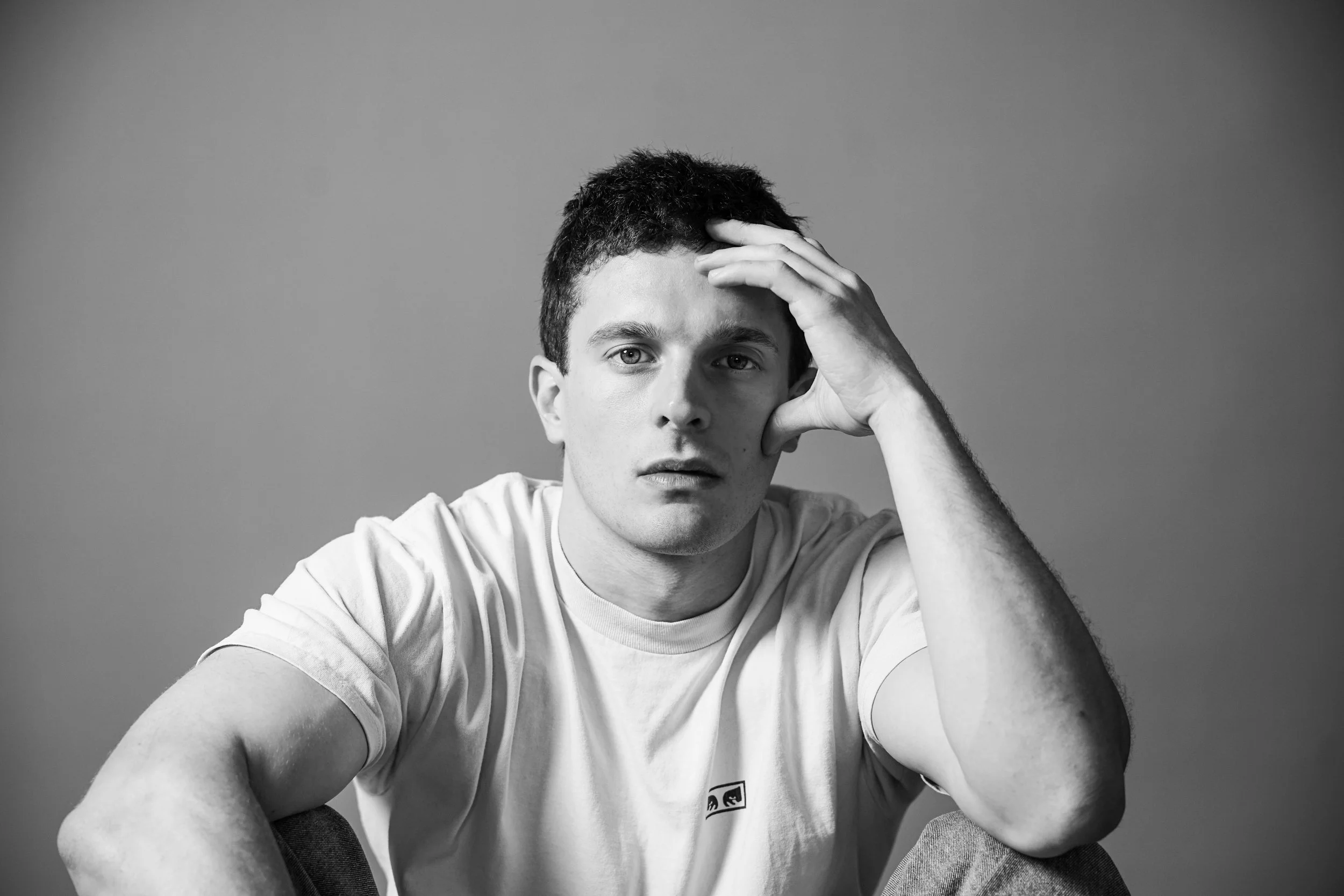 Ragazzo seduto con espressione pensierosa, mano sulla testa, sfondo grigio, in bianco e nero.