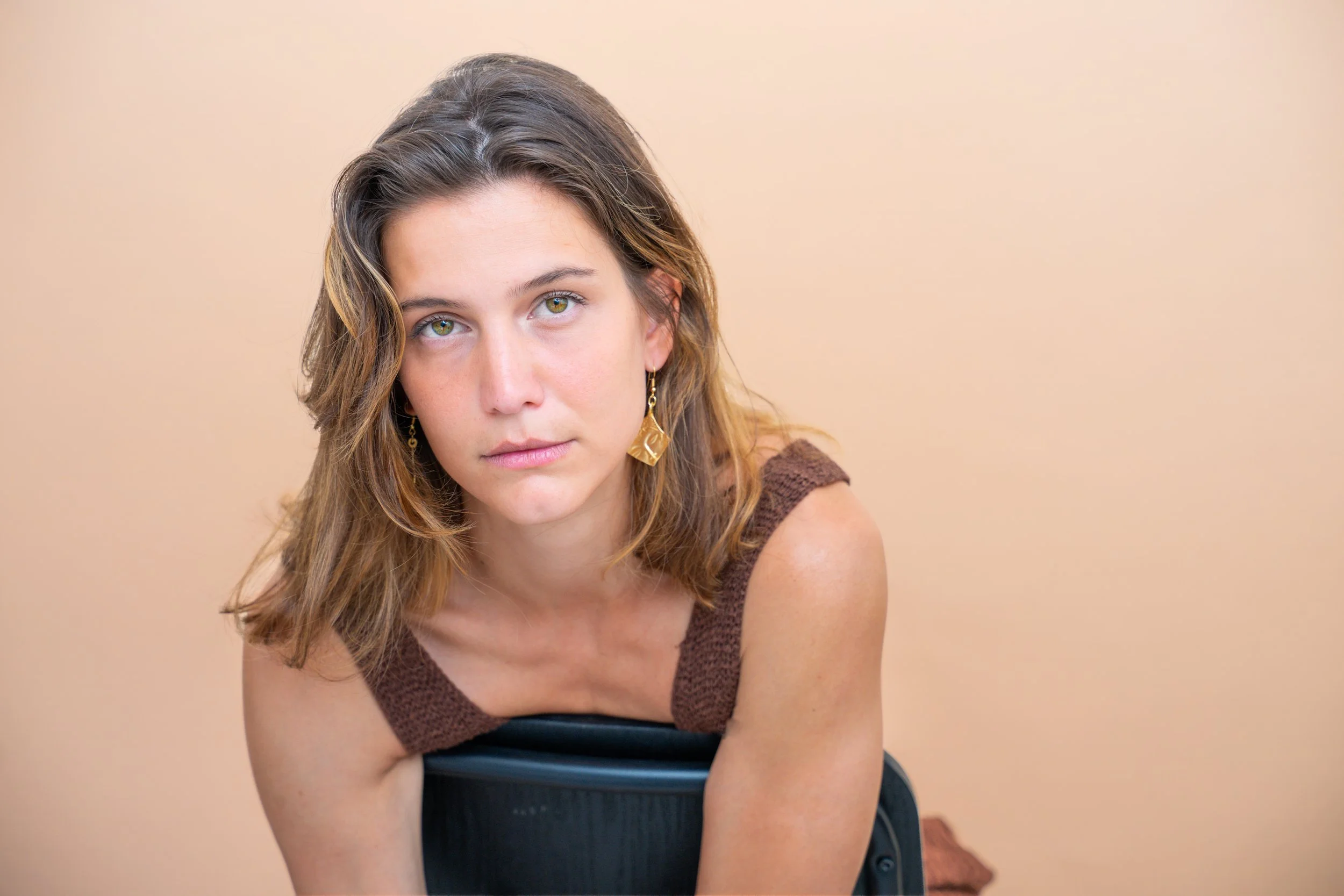 Ragazza dai capelli castani riccioli, occhi verdi, con orecchini dorati, vestita con top marrone e nero, seduta su una sedia contro uno sfondo beige.