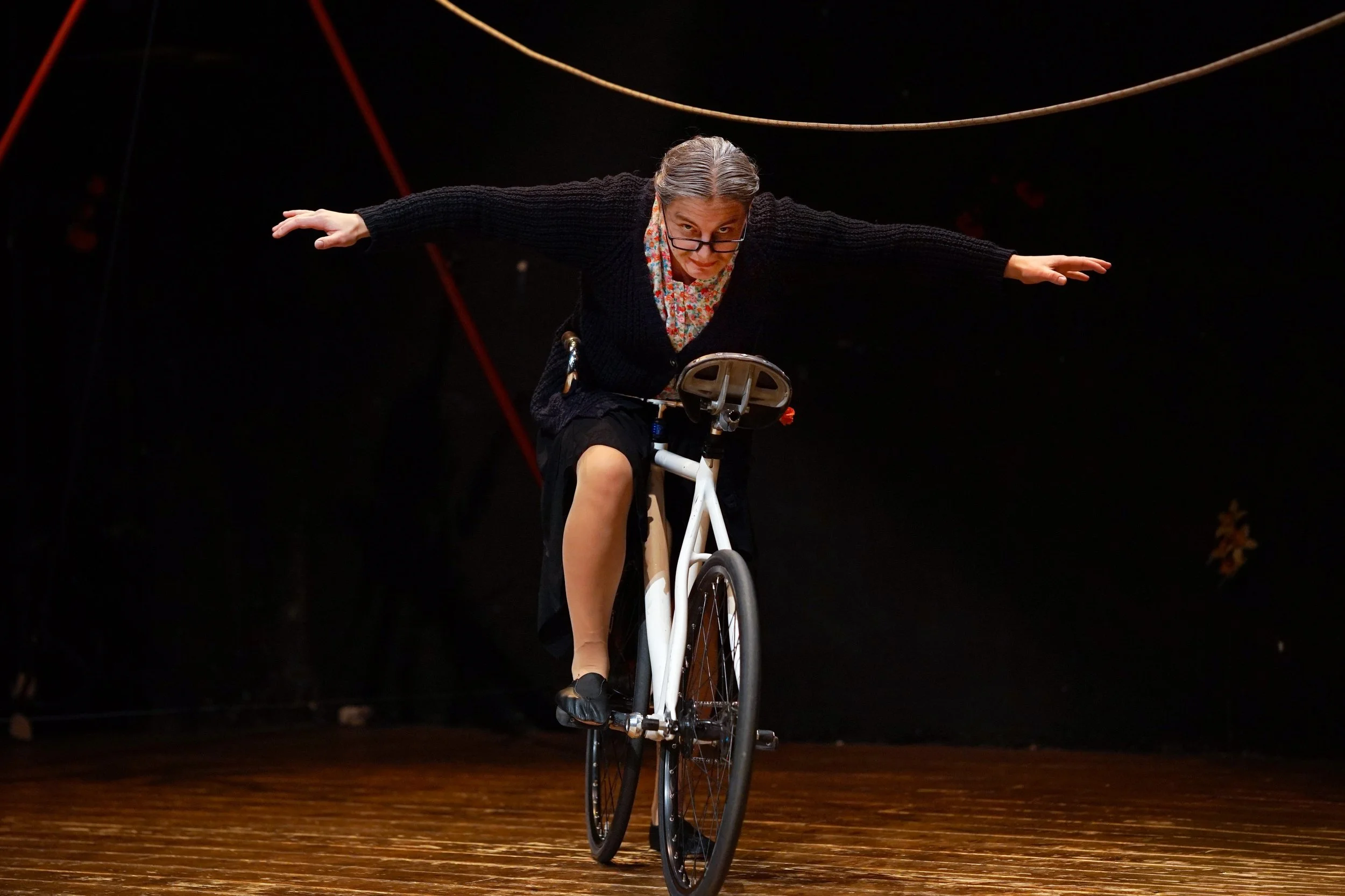 Una donna vestita casualmente con pantaloncini e maglione nero sta facendo l'equilibrista su una bicicletta statica su un palco di legno, con la mani tese verso i lati per mantenere l'equilibrio, durante una performance teatrale o circense, con sfond
