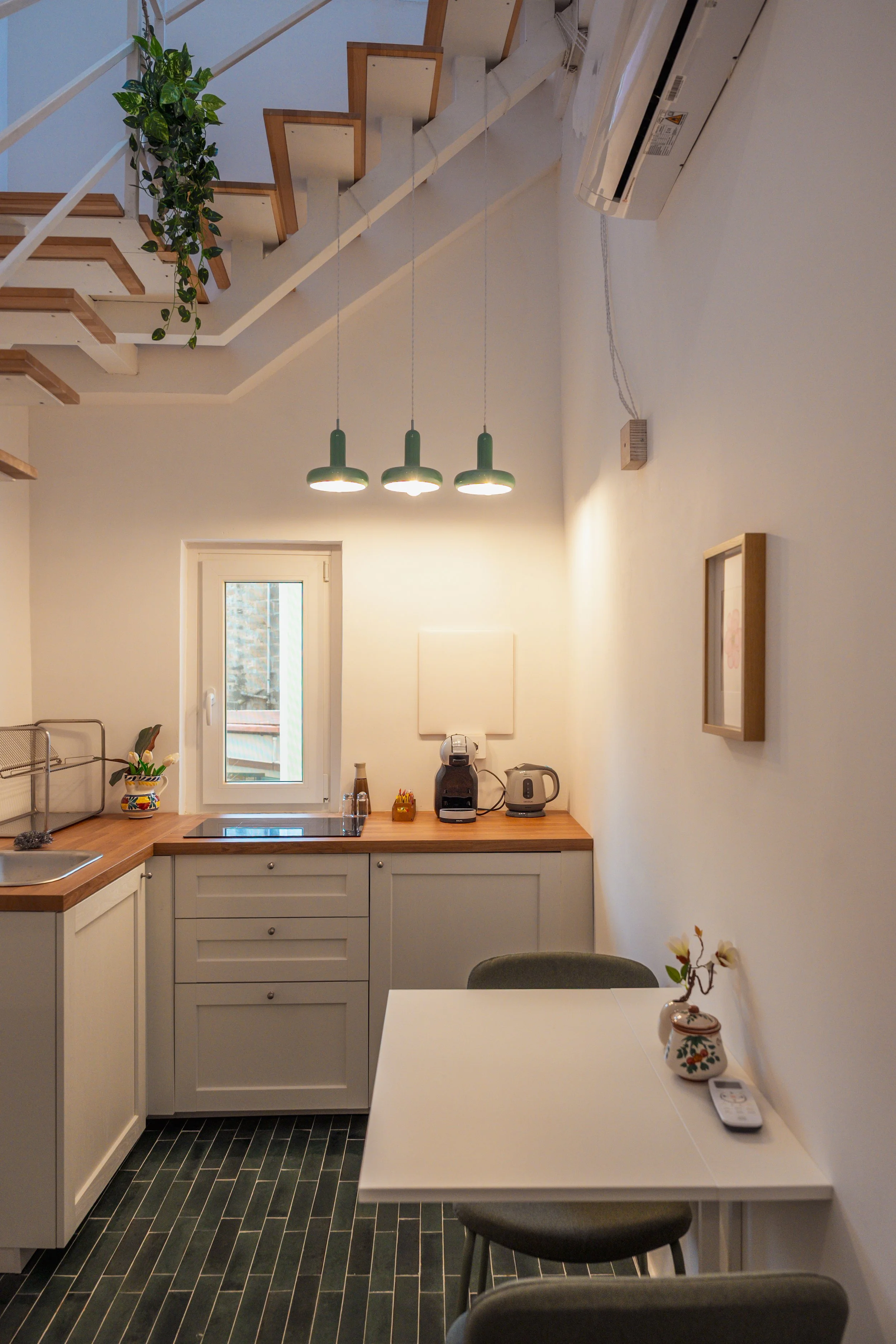 Piccolo angolo cottura con cucina bianca, piani in legno, finestrella, lampade sospese verdi, tavolino con sedie, decorazioni e piante.