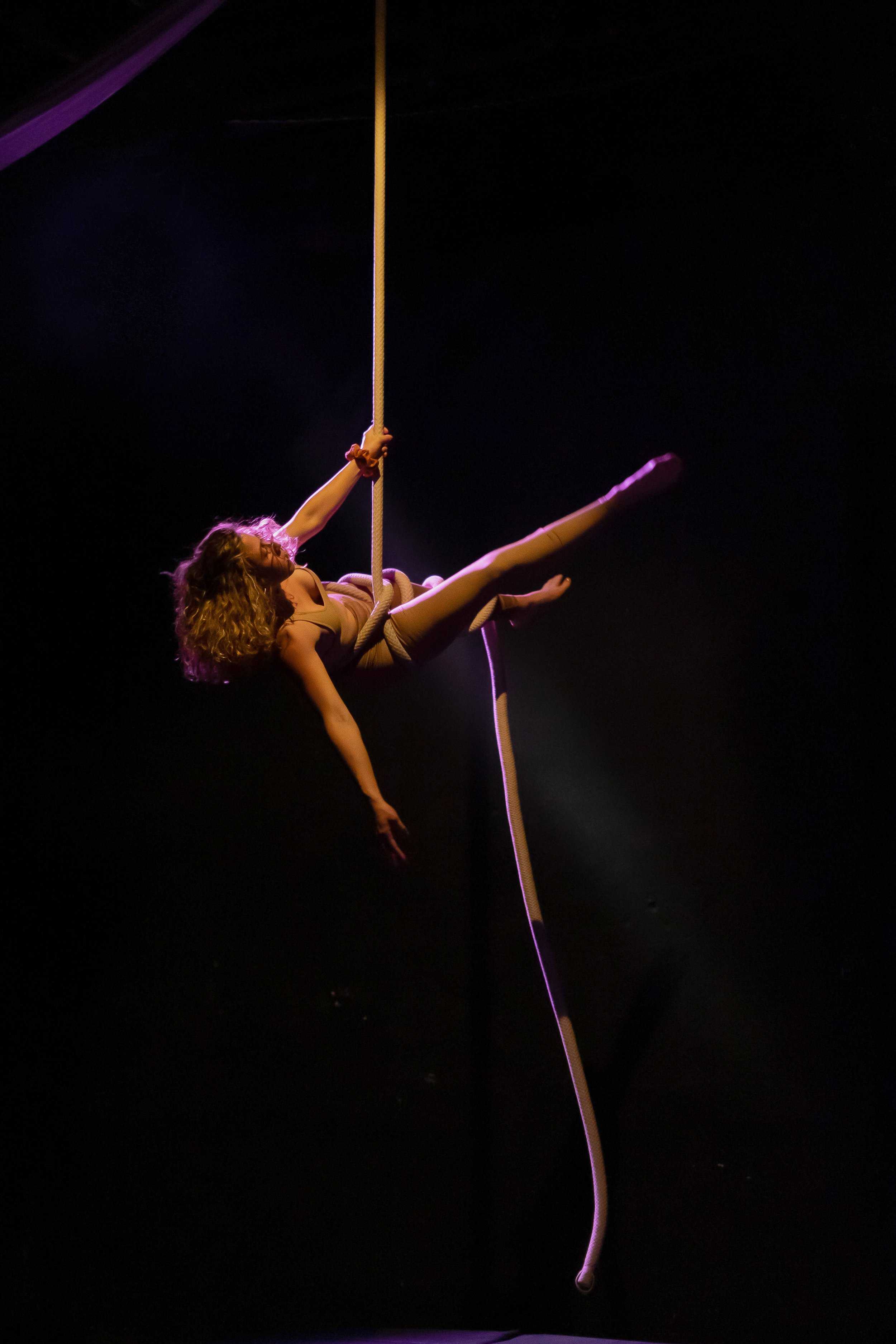 Una donna si esibisce in una performance di pole dance su un'asta, sospesa nell'aria contro uno sfondo nero, illuminata da luci soffuse.
