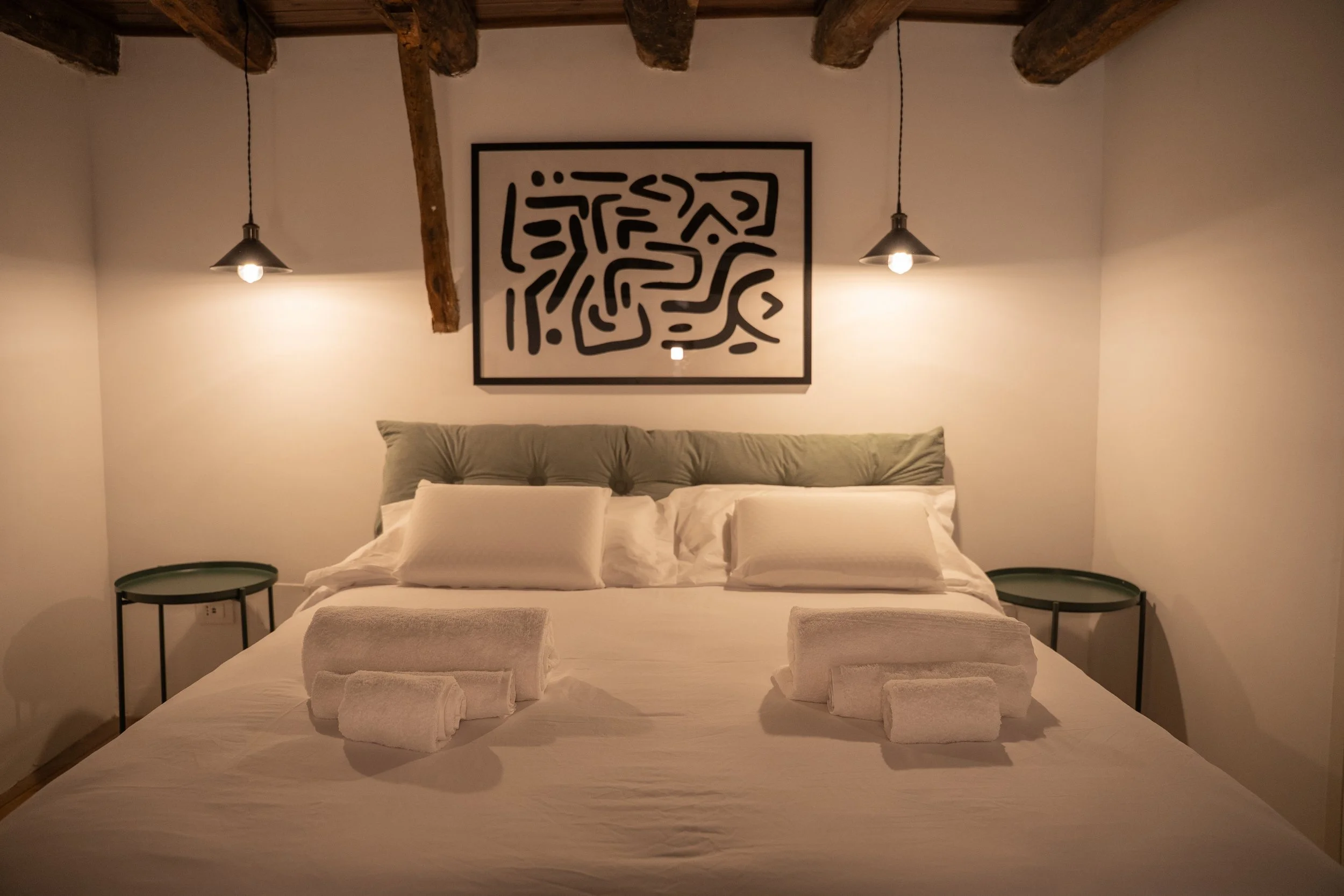 Letto matrimoniale in una stanza con decorazioni minimaliste, due lampade appese, quadri astratti e panni puliti appoggiati sul letto.