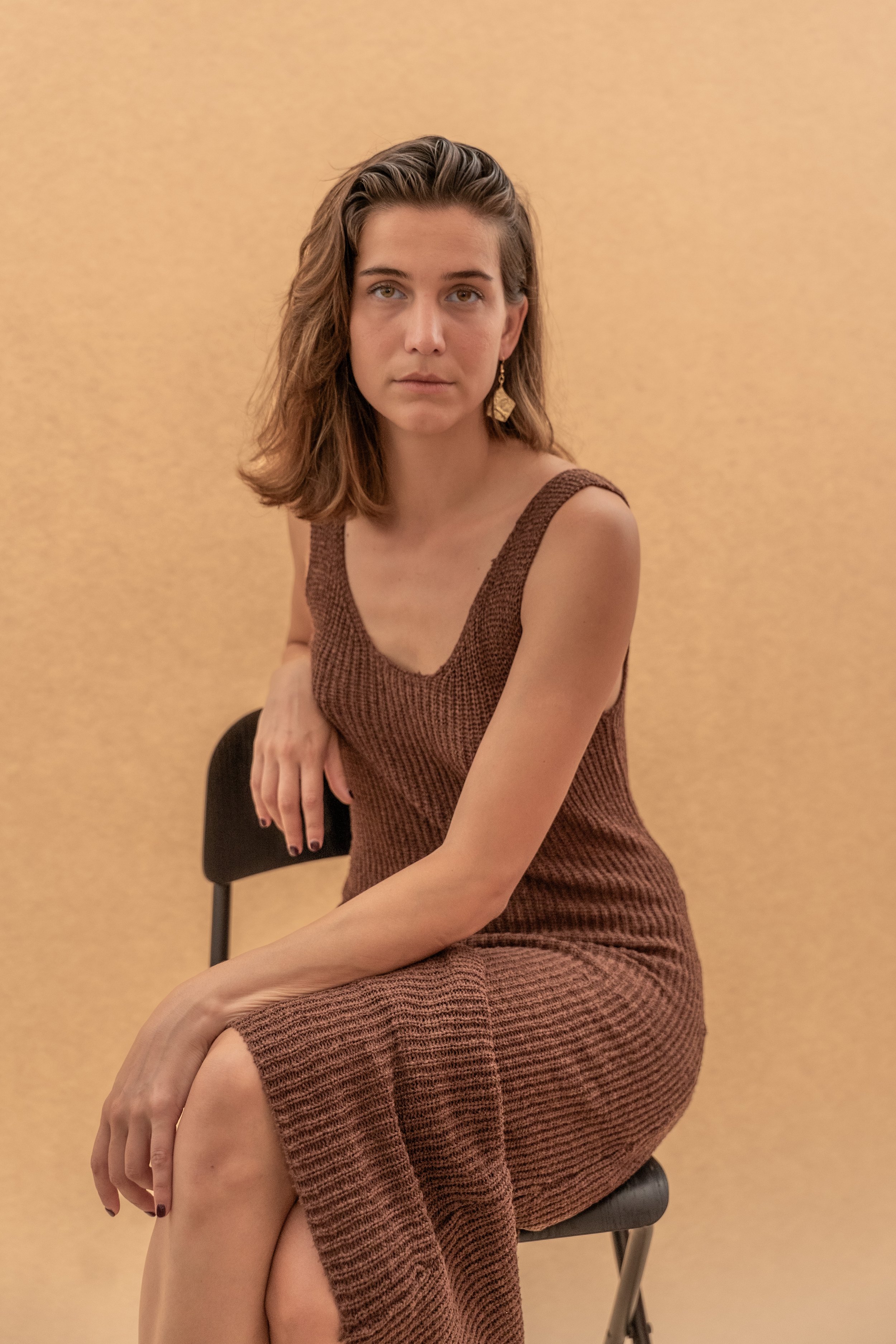 Una donna seduta su una sedia contro uno sfondo beige, indossa un vestito di maglia marrone a righe, con capelli castani ondulati e orecchini grandi dorati.