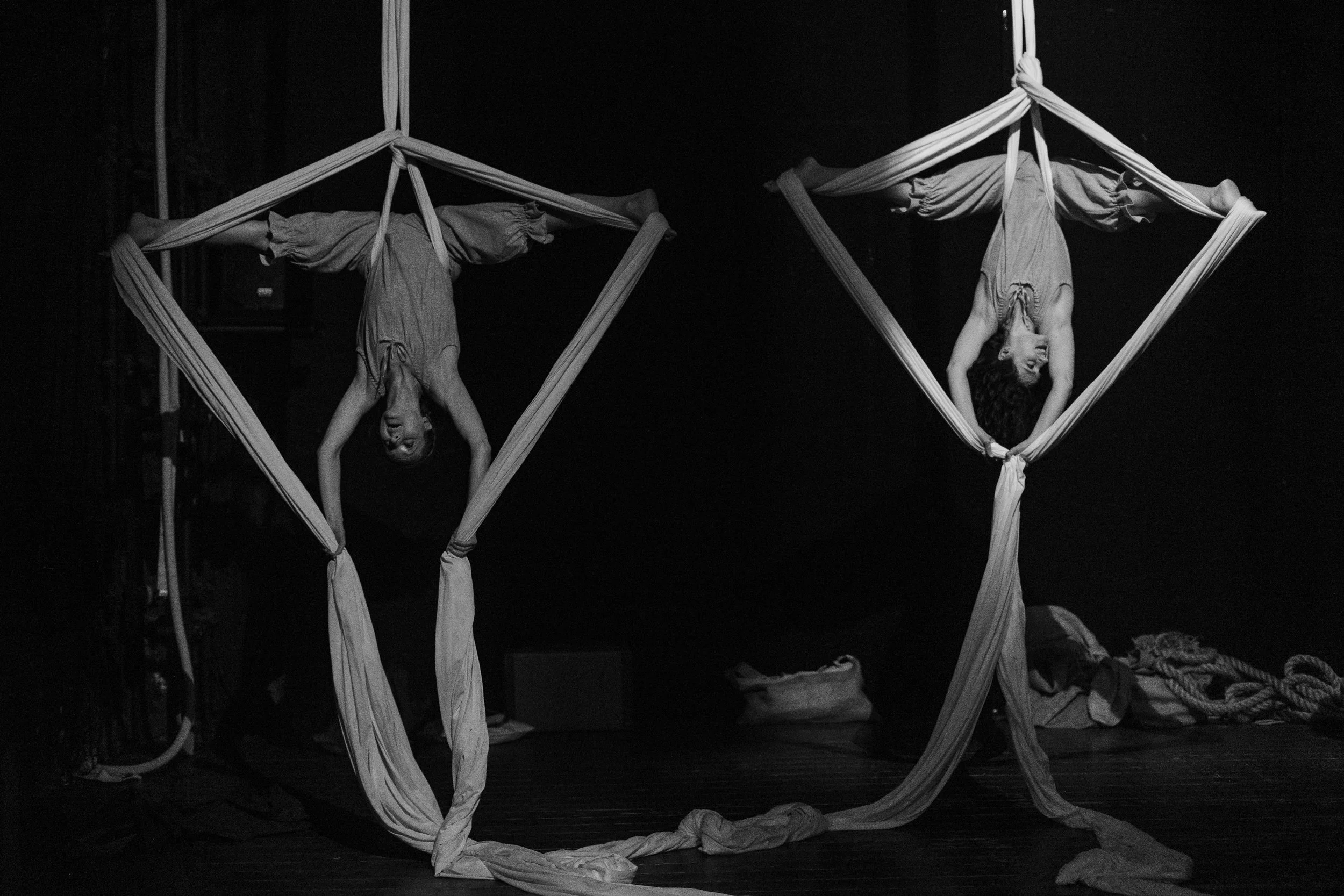 Due persone si esercitano in acrobata con tessuti da circo in uno spazio nero, una donna è capovolta con le gambe appoggiate su un tessuto, l'altra è capovolta con le ginocchia piegate e si tiene con le mani al tessuto.