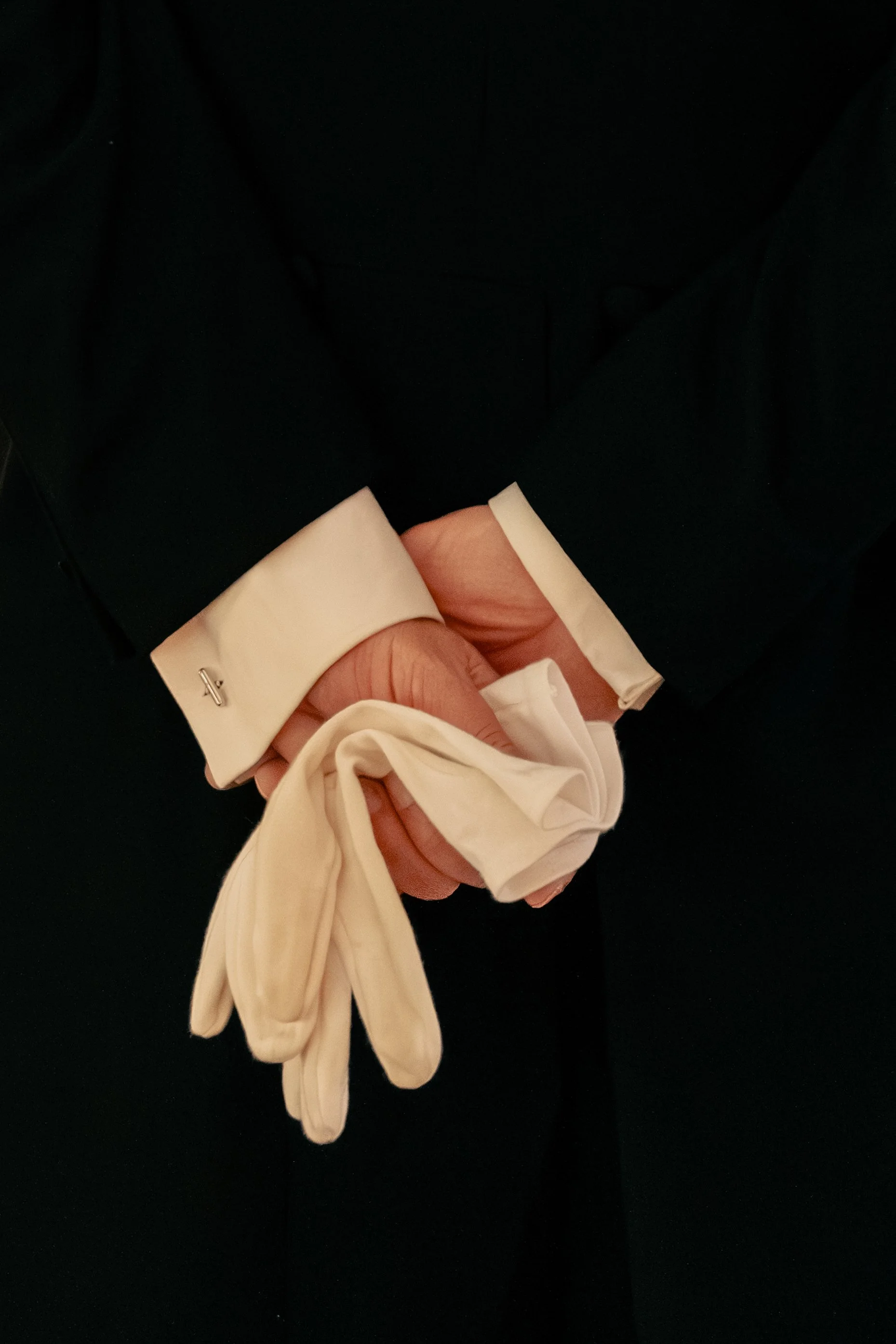 Una persona indossa un abito scuro con polsini bianchi, che tiene in mano dei guanti in lattice beige.