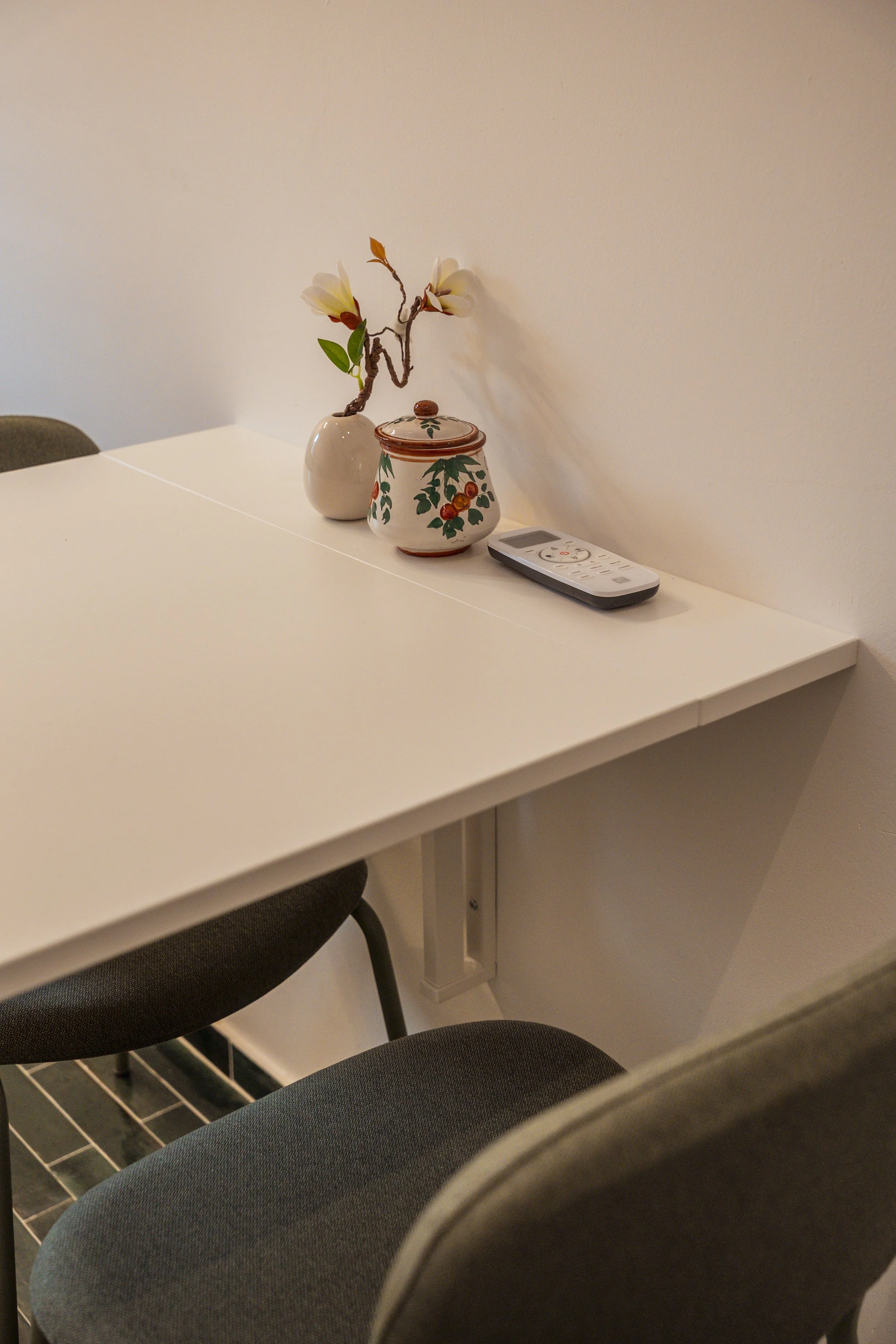 Tavolo bianco con una sedia grigia, decorato con un vaso bianco con un ramo di fiori gialli, una scatola decorativa con motivi floreali e una telecomando su un muro bianco.