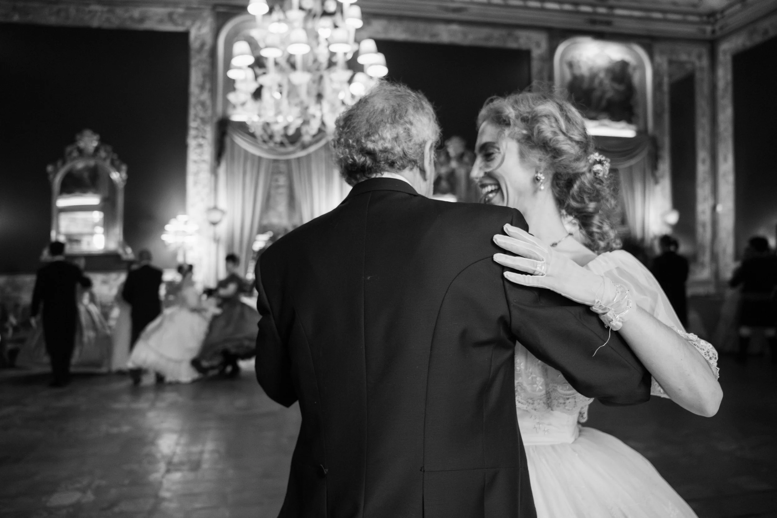 Un matrimonio che danza nel salone di un matrimonio elegante, con il marito vestito in nero e la sposa in abito bianco, sorridenti e felici.
