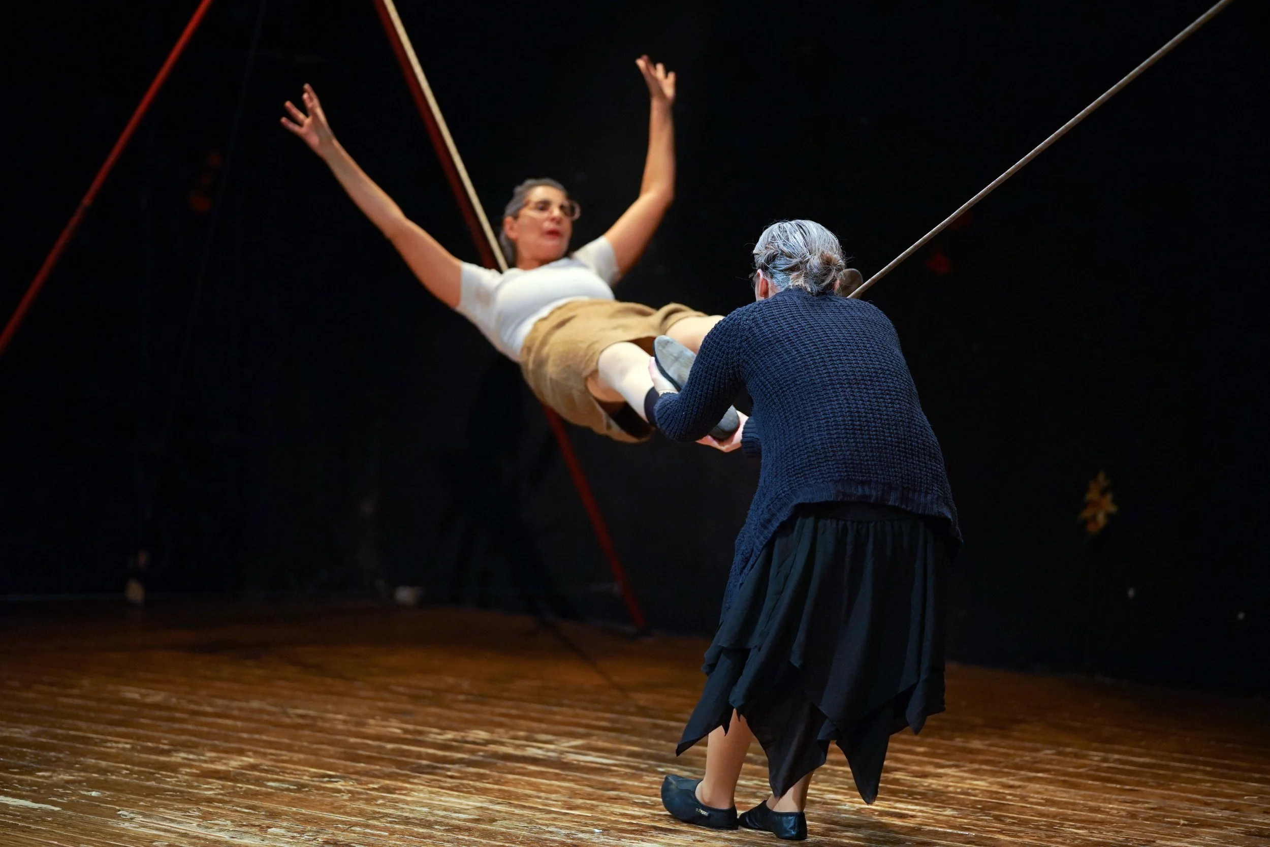 Due donne praticano un'esibizione di teatro o danza, con una donna che viene sollevata o sospesa in aria mentre un'altra la sostiene.