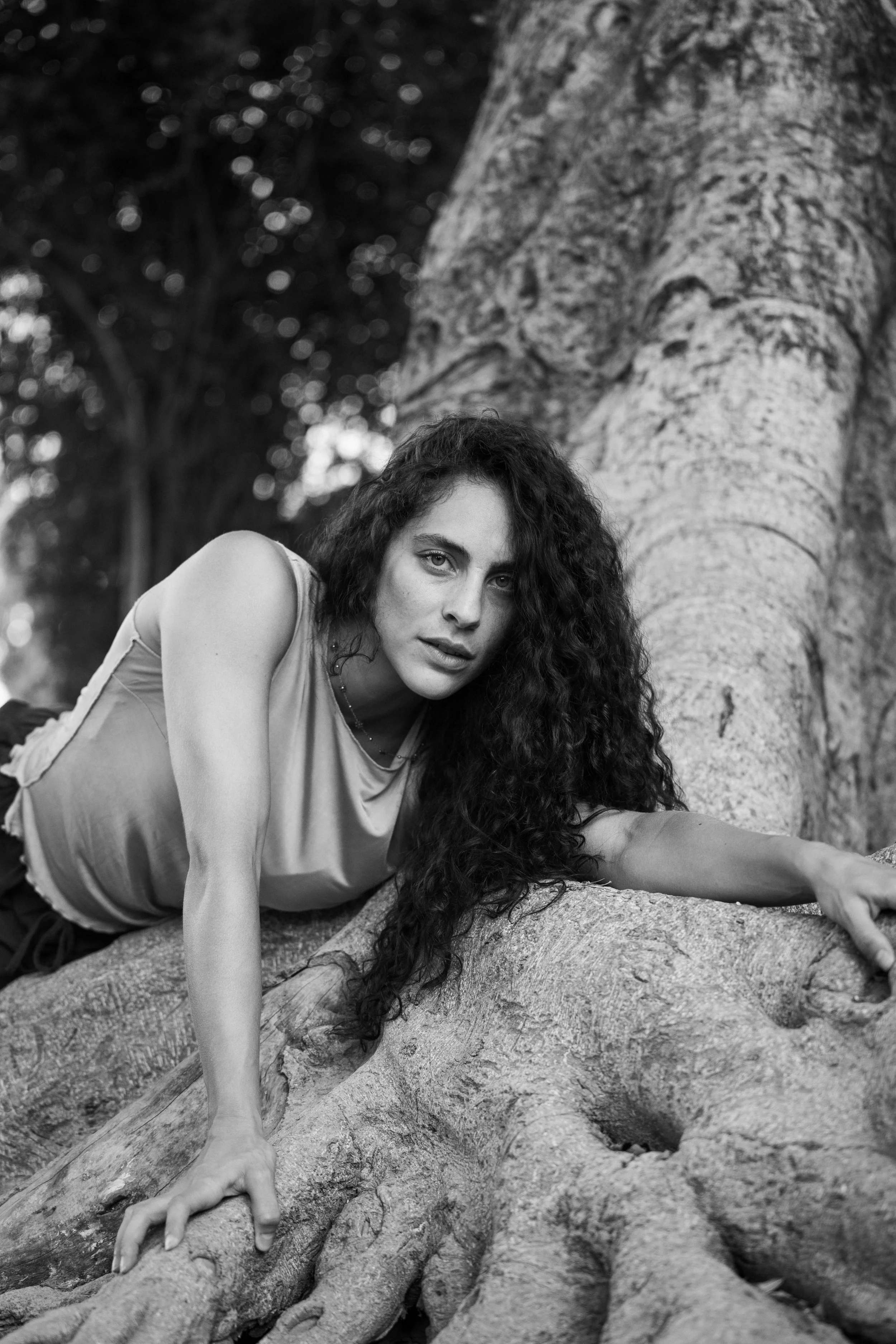 Una donna con capelli ricci stretti si sdraia su un tronco di albero, in posa artistica, tra alberi e foglie in uno scenario naturale.
