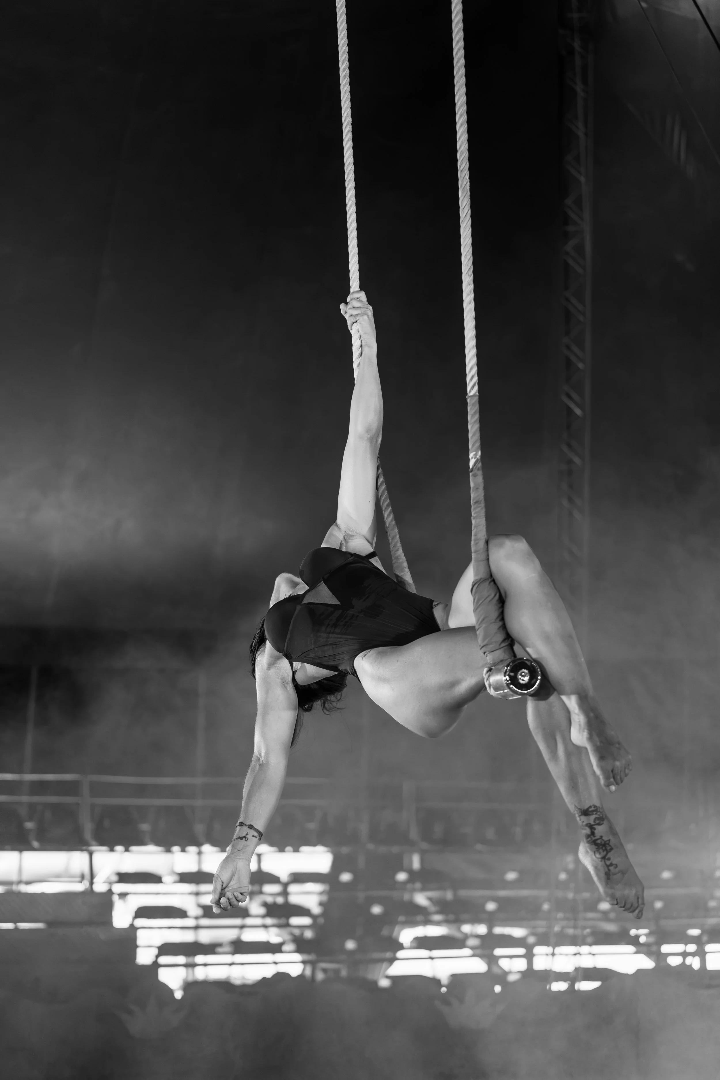 Una donna si trova in una palestra, appesa a due corde nello stile di un acrobata o di una performer di spettacoli acrobatici, con un'espressione concentrata. L'immagine è in bianco e nero.