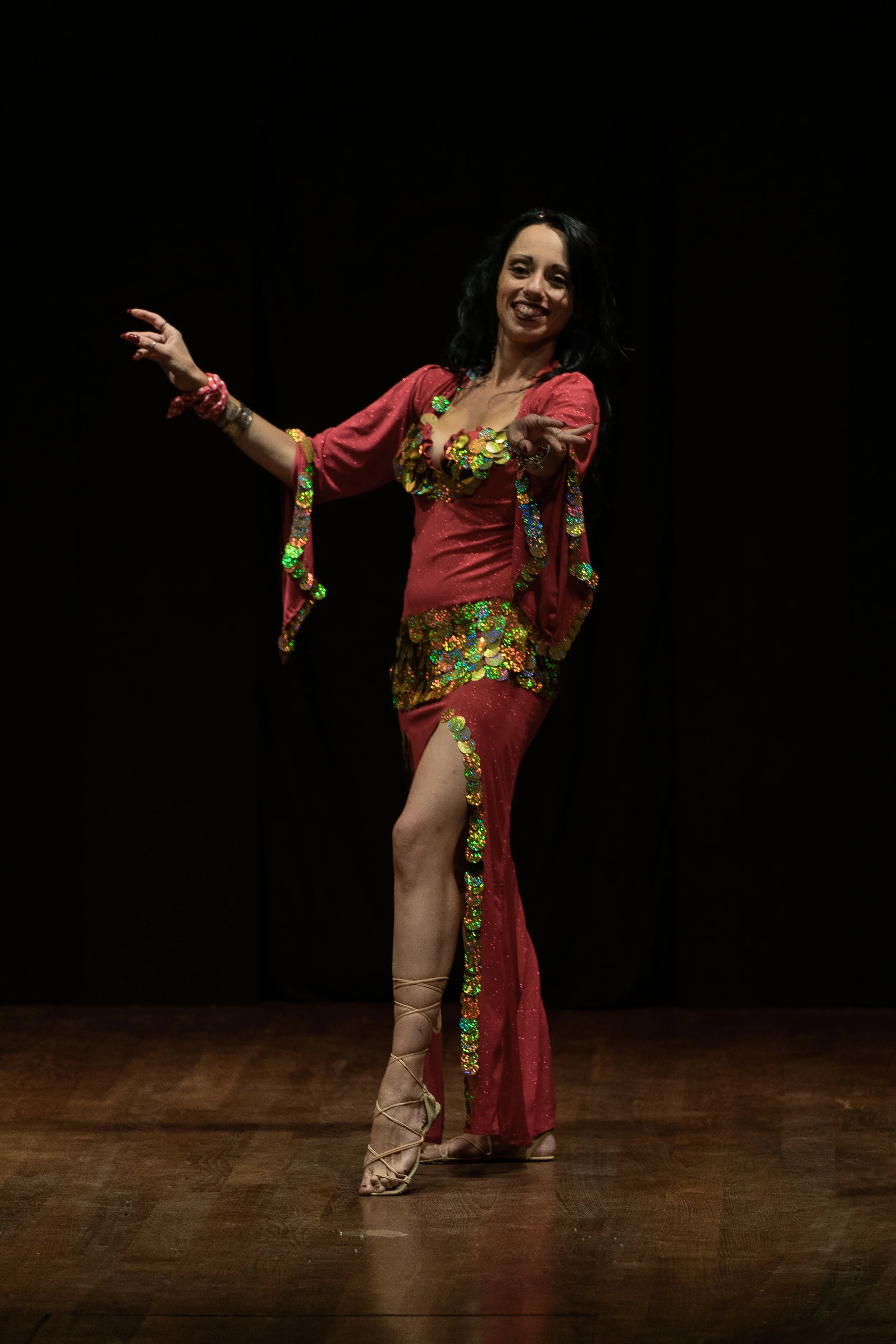 Donna che danza folkloristica su palco con abito rosso decorato con paillettes, con una gamba in posizione di danza e braccia aperte, sfondo nero.