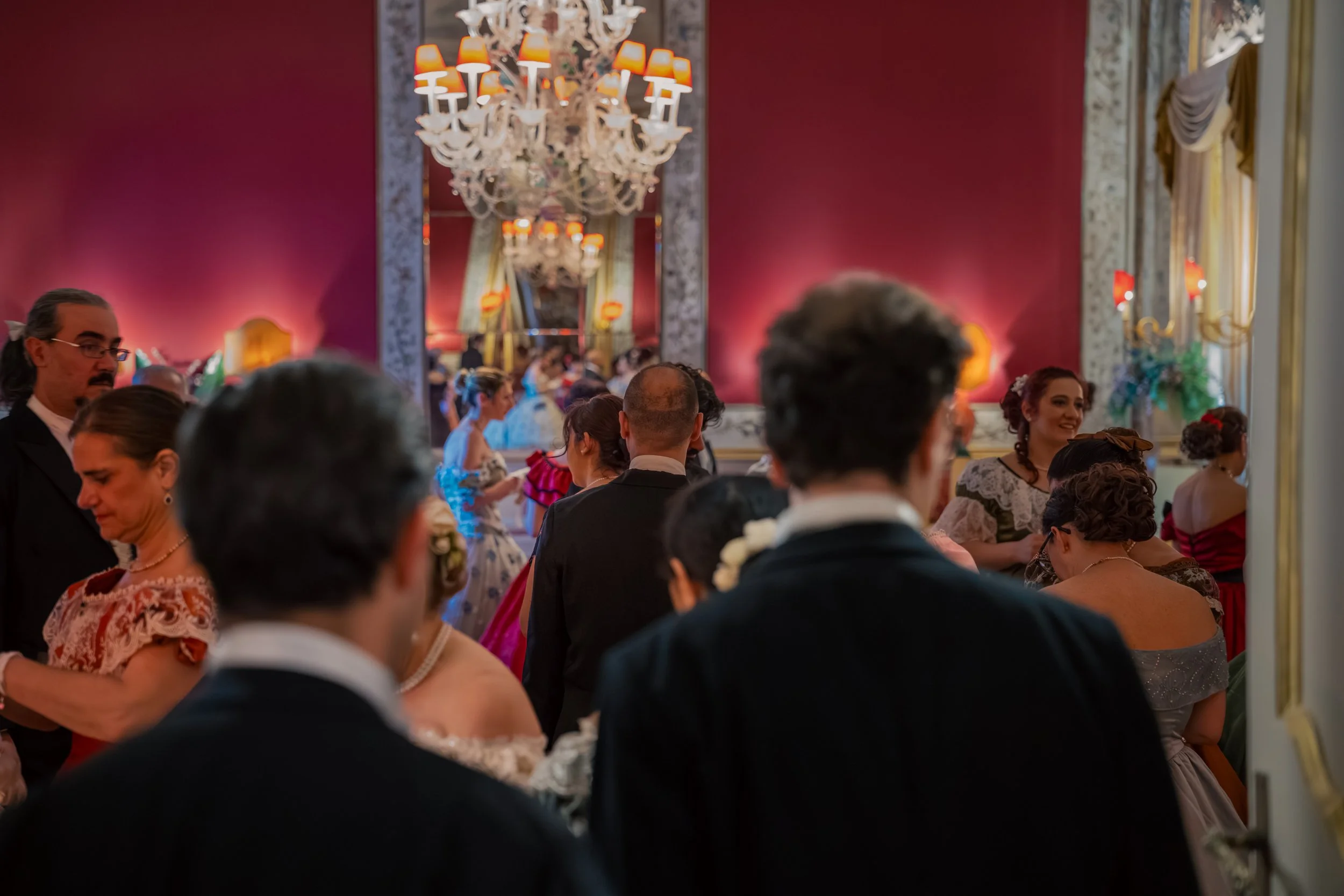 Persone vestite elegantemente in un salone decorato con pareti rosse, grandi specchi e lampadari, durante un evento sociale.