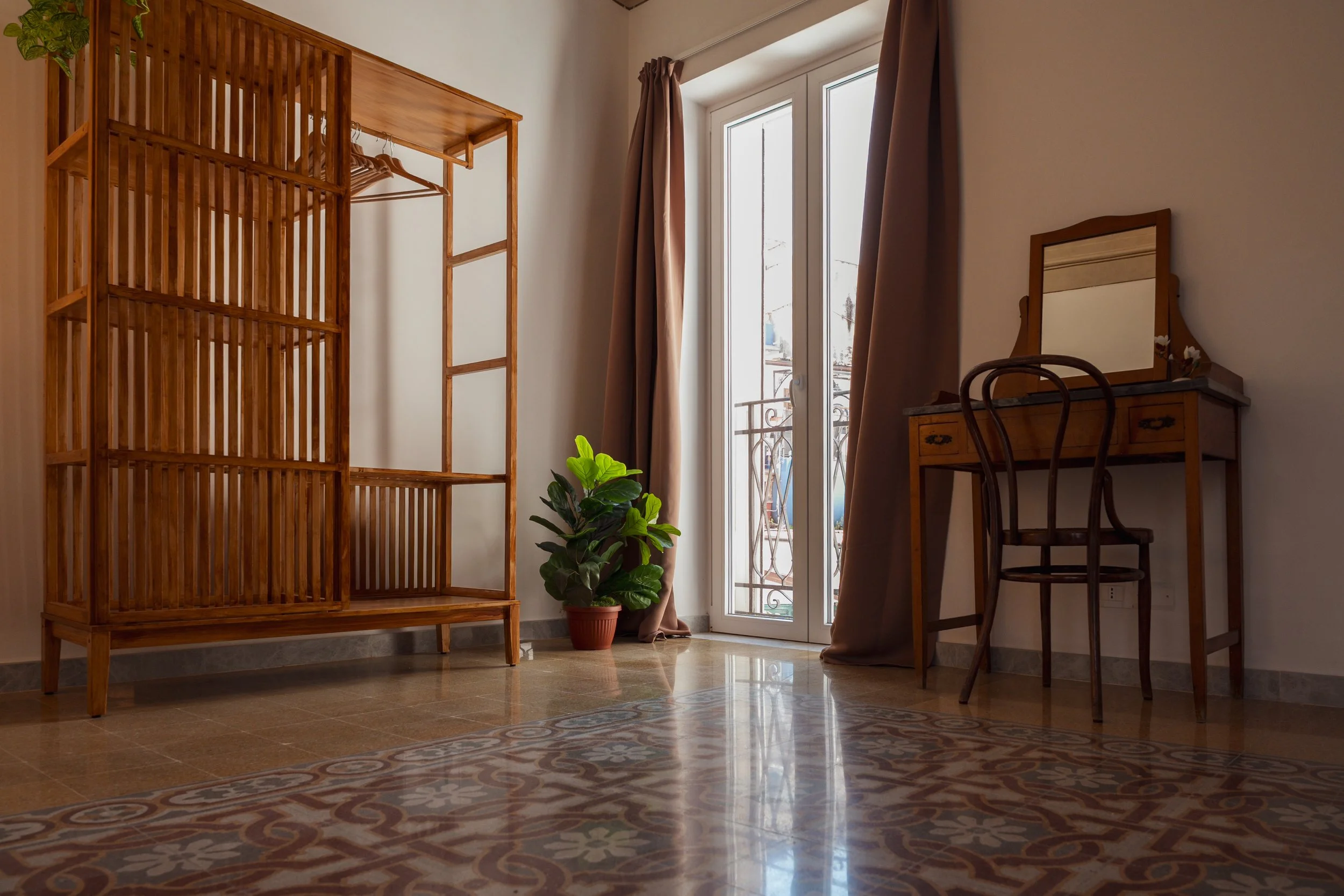 Interno di una stanza con porta finestra, armadio in legno con mensole e gruccia, tavolo e sedia in stile classico, pianta in vaso, tappeto con motivo floreale, tende marroni.