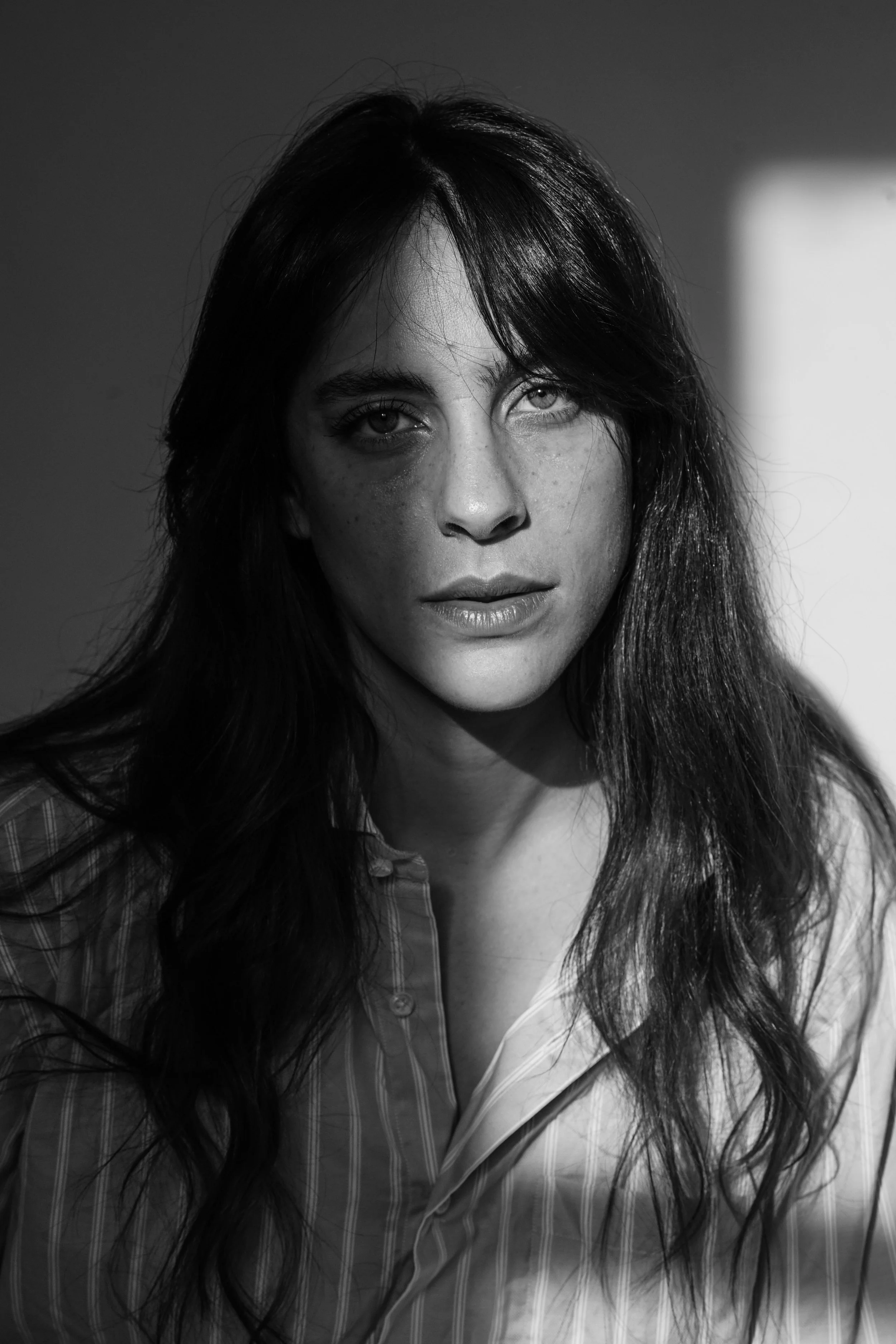 Ritratto in bianco e nero di una donna con capelli lunghi e scuri, che indossa una camicia a righe. La luce crea un'ombra sulla parete dietro di lei.