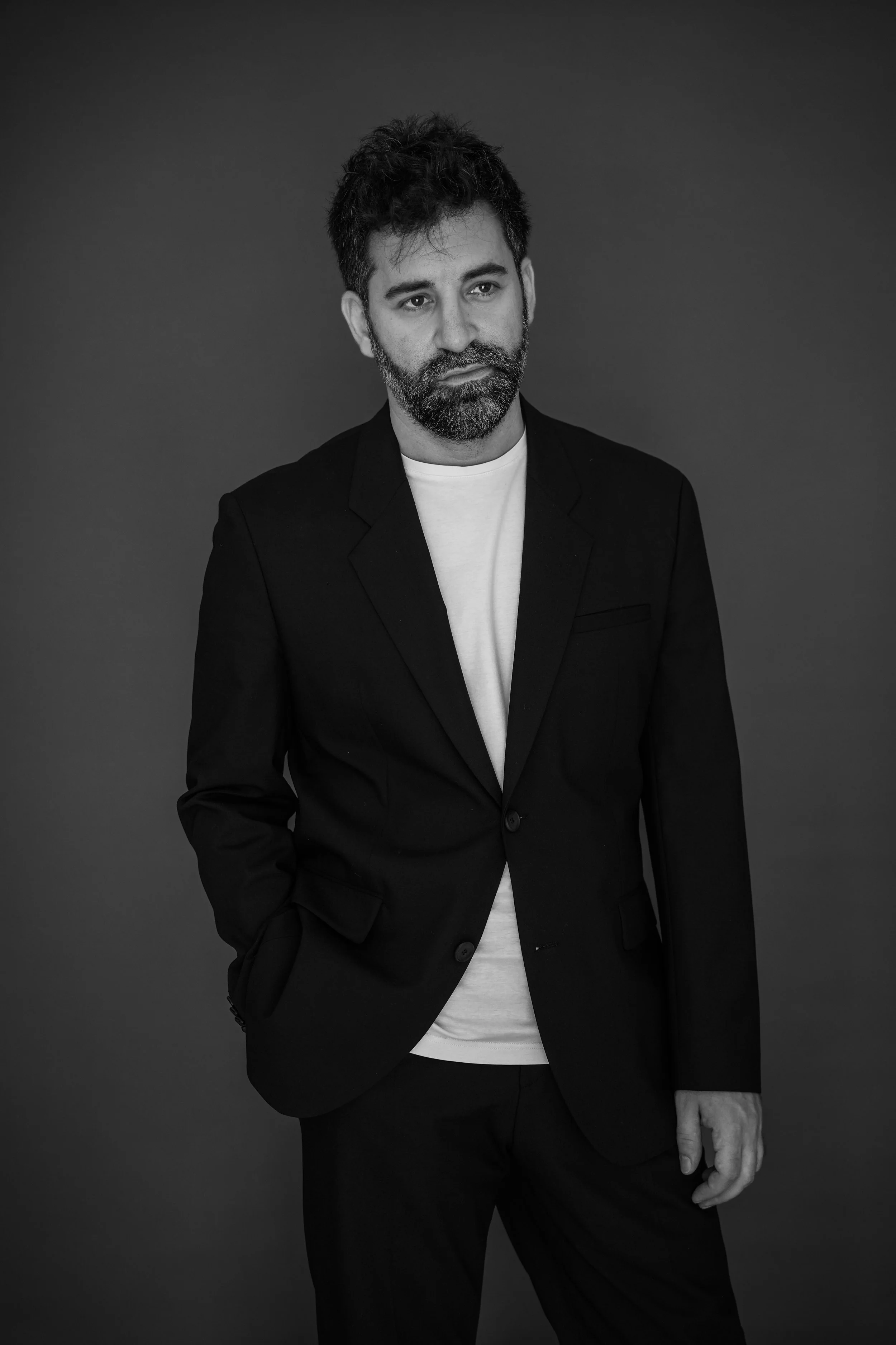 Ritratto in bianco e nero di un uomo con barba e capelli ricci, indossa un blazer nero sopra una maglietta bianca, posando con una mano in tasca contro uno sfondo scuro.