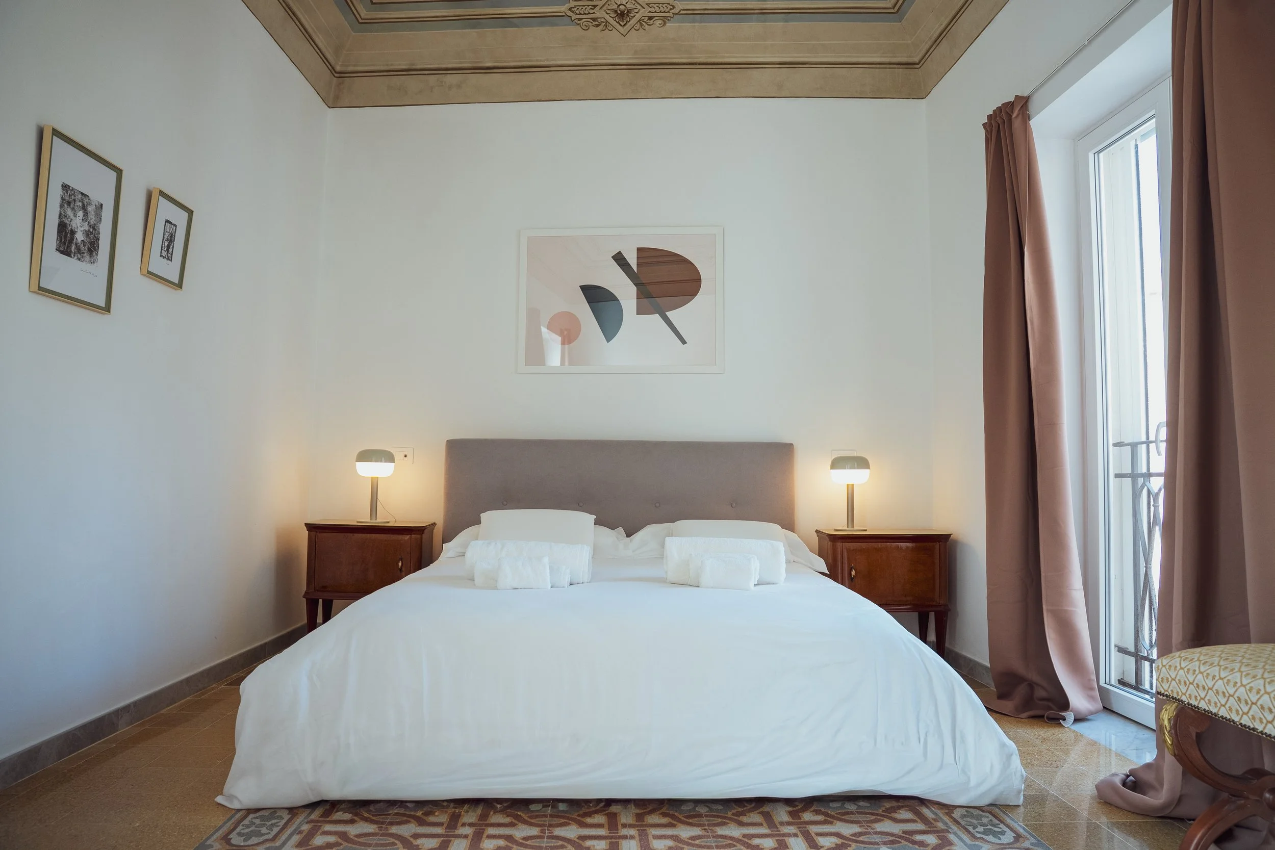 Camera da letto con letto matrimoniale, testiera imbottita, lenzuola bianche, due comodini con lampade, quadro astratto sopra il letto, pareti bianche, finestra con tende rosa, pavimento in piastrelle, parte di poltrona o sedia con rivestimento dorat