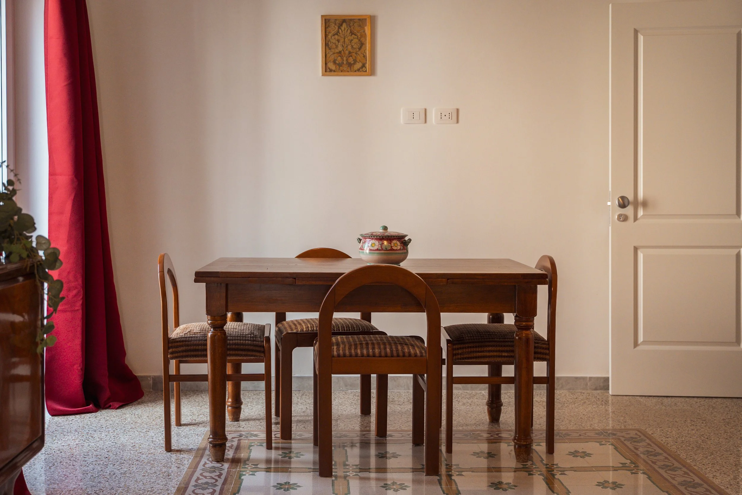 Una stanza con tavolo e sedie in legno, piastrelle decorate, pareti bianche, quadro appeso, porta bianca e tende rosse.