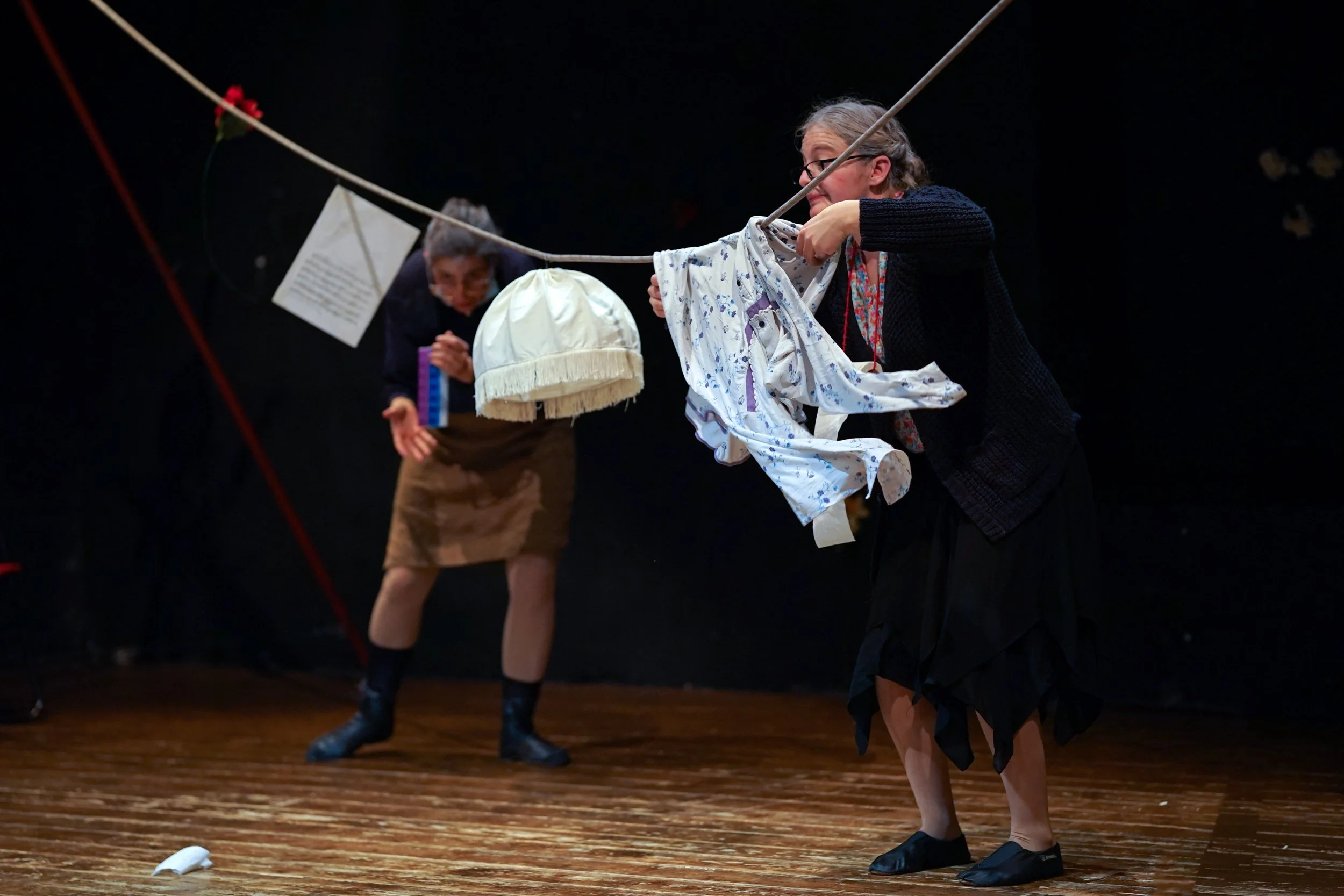 Due donne anziane interpretano una scena teatrale con abiti e oggetti di scena, una tiene in mano dei vestiti e l'altra si inarca verso un filo con bambole e un cappello pendenti, sul palcoscenico con pavimento di legno e sfondo nero.
