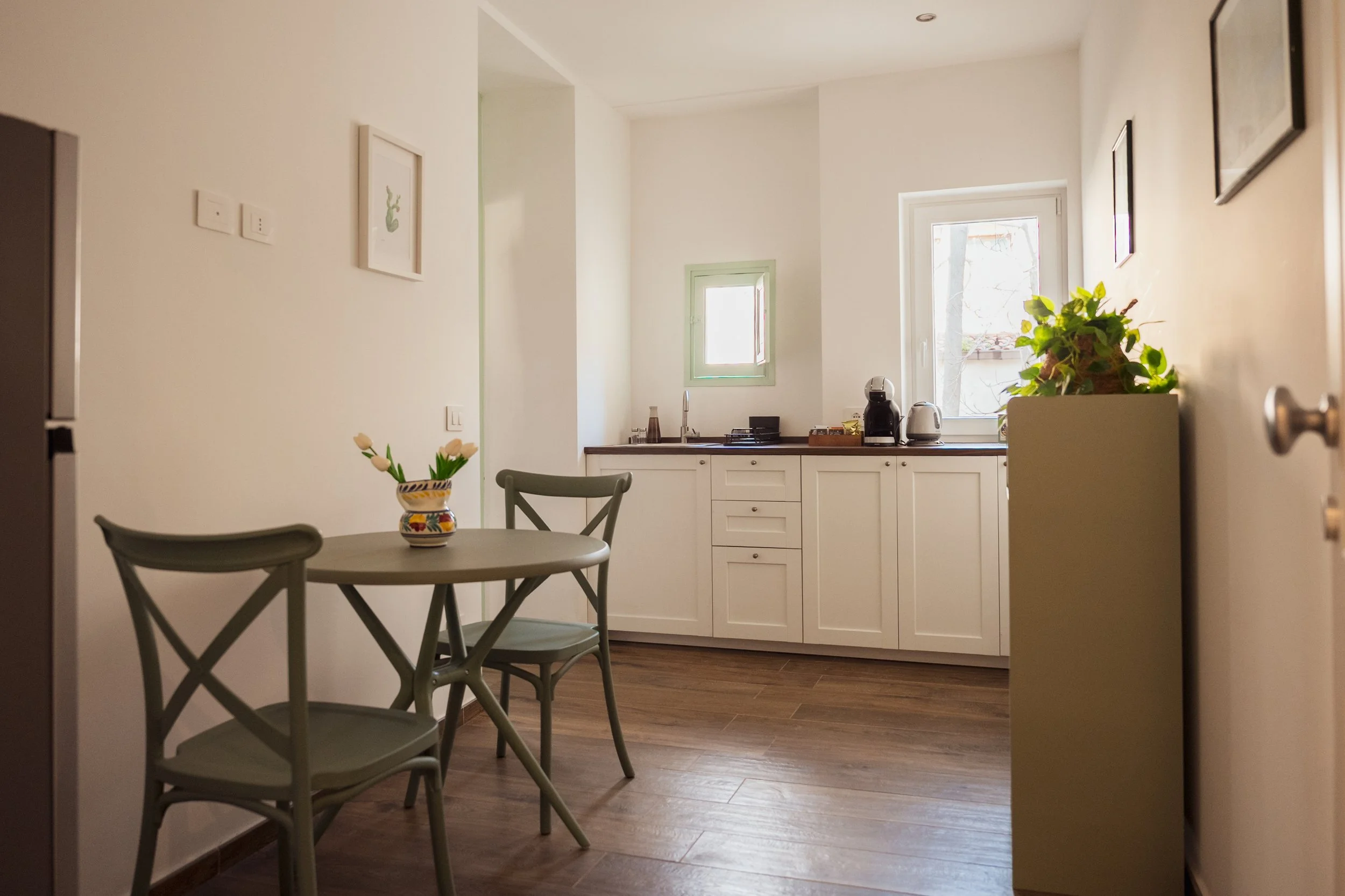 Angolo cottura con mobili bianchi, due finestre, tavolo con due sedie e una pianta sulla credenza. Quadro decorativo alle pareti e pianta decorativa sul mobile.