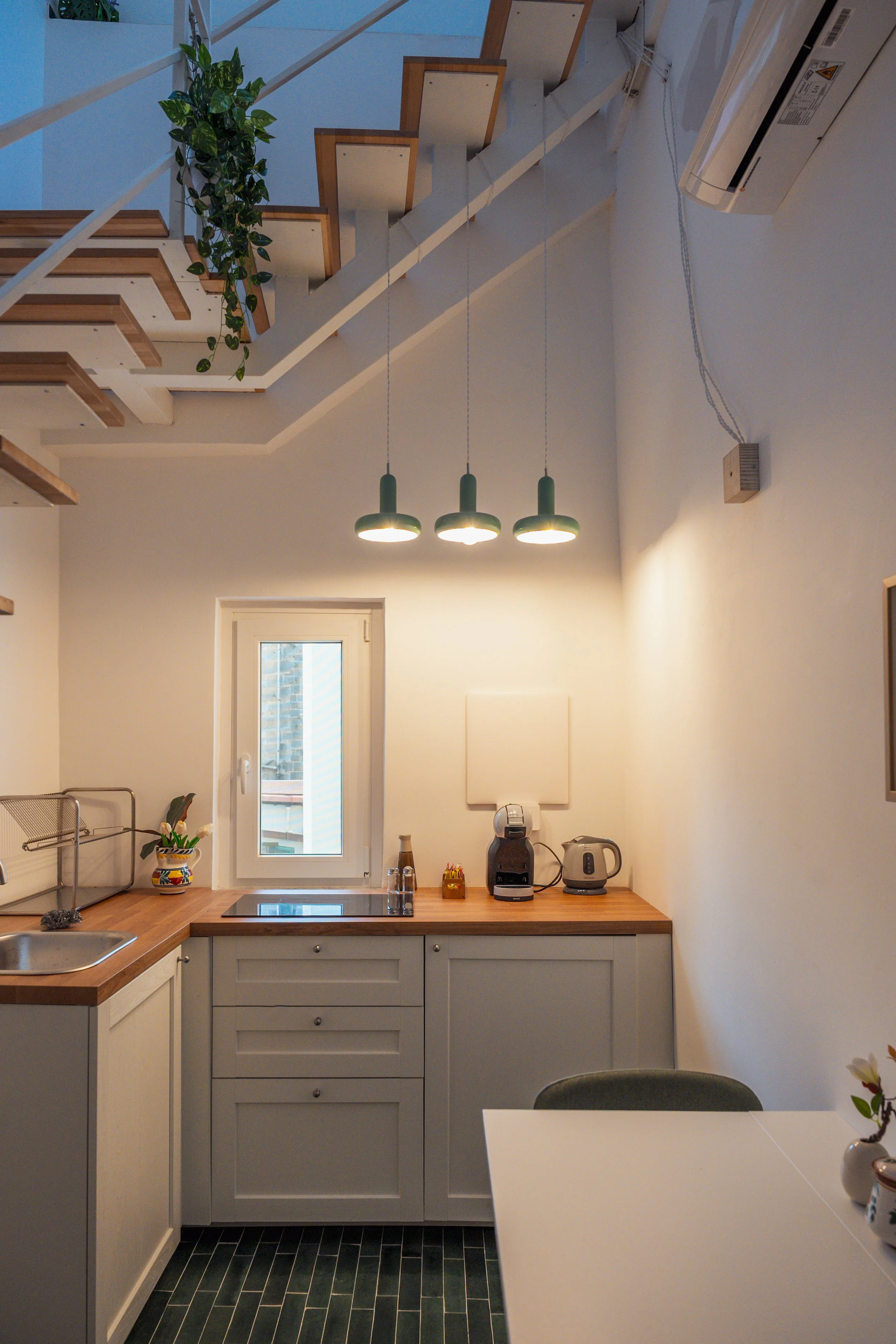Cucina moderna con armadietti bianchi, piano di lavoro in legno, lampade verdi sospese, una finestra, alcuni elettrodomestici come macchina da caffè e termosifone, e un tavolo con sedie.