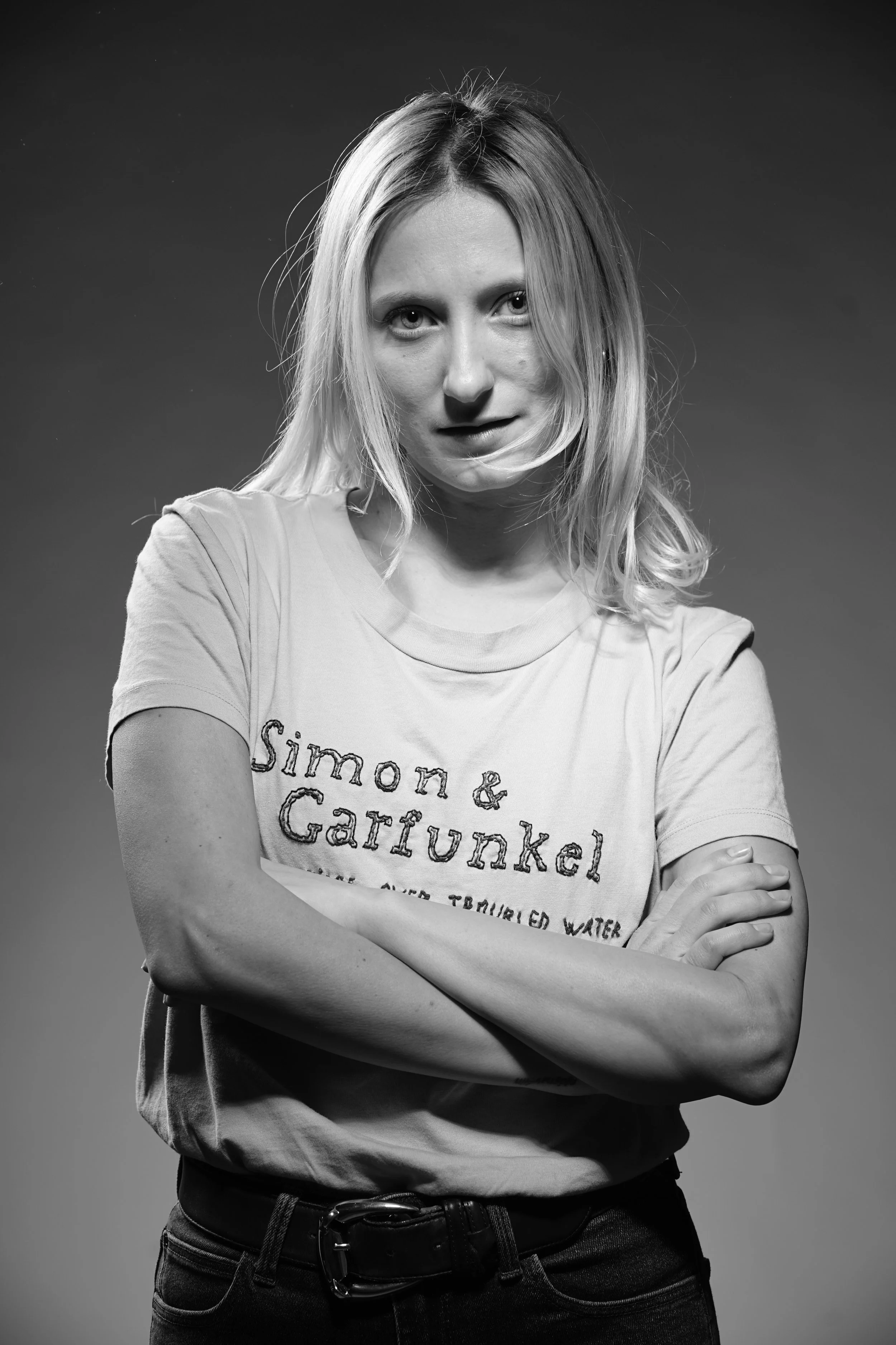Ragazza con capelli biondi, vestita con una maglietta con scritta 'Simone & Garrunkel', che si incrocia con le braccia e guarda verso la fotocamera in un'immagine in bianco e nero.