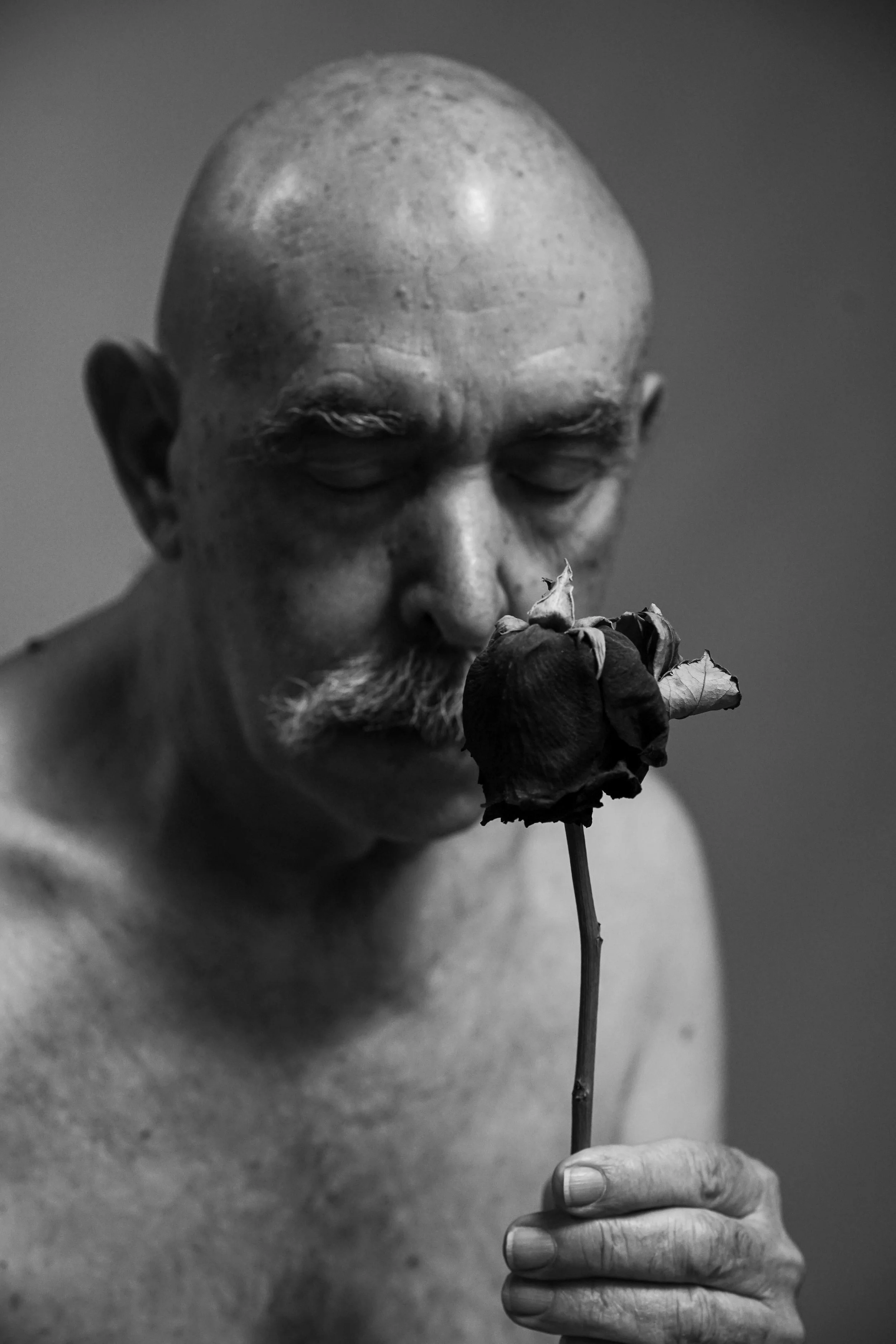 Ritratto in bianco e nero di un uomo anziano senza maglietta con un baffo e capelli rasati che tiene una rosa appassita vicino al volto. L'uomo ha gli occhi chiusi.
