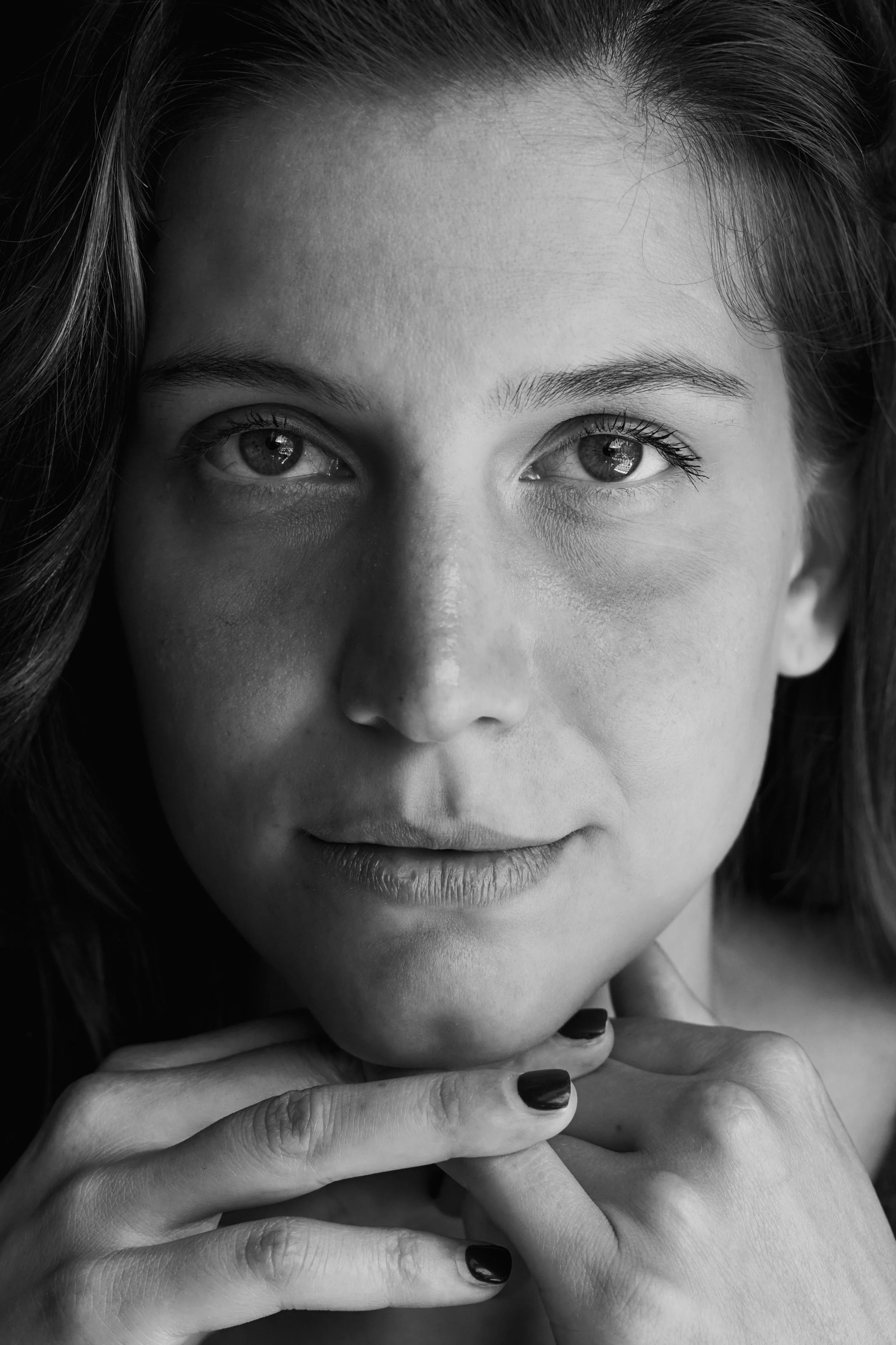Ritratto in bianco e nero di una donna che guarda verso l'obiettivo, con le mani sotto il chin, capelli mossi e occhi espressivi.