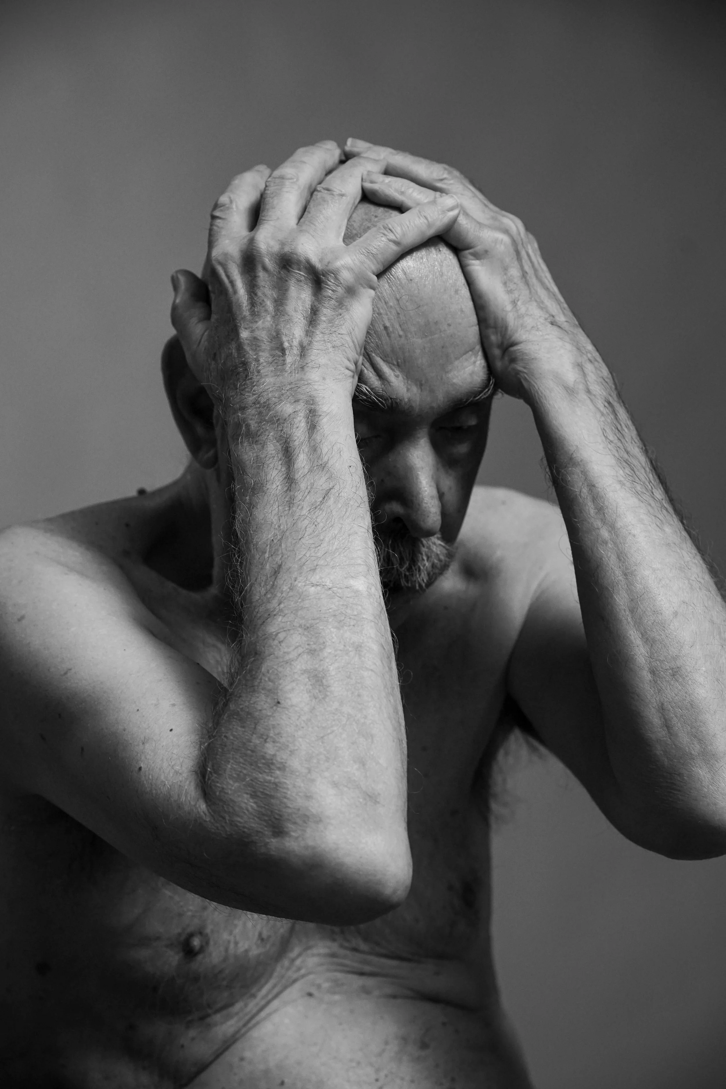 Uomo anziano senza maglietta, con capelli e barba grigia, che si tiene la testa tra le mani, esprimendo ansia o stress.
