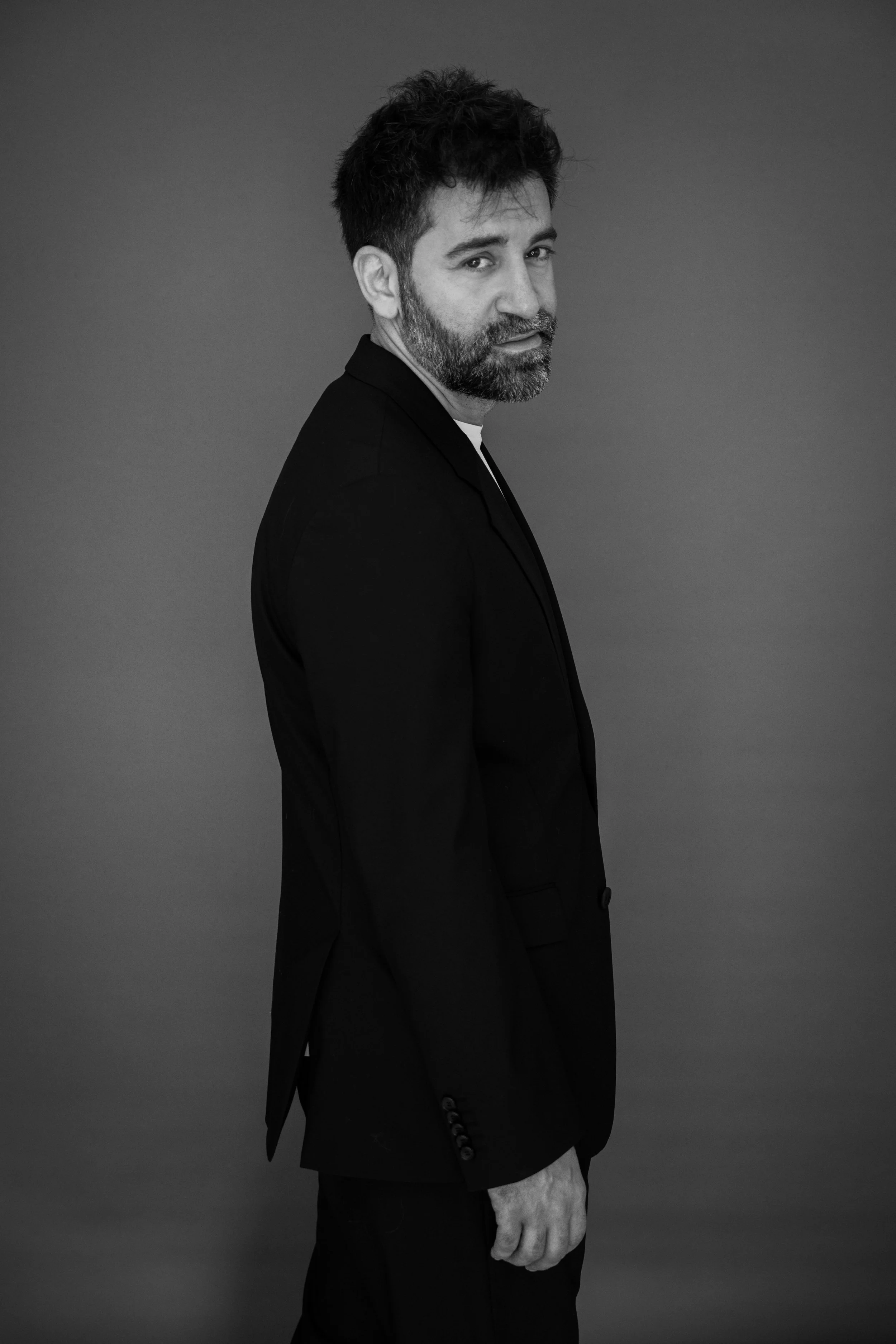 Ritratto in bianco e nero di un uomo con barba e capelli corti, vestito con giacca nera, che guarda di lato su uno sfondo uniforme grigio.