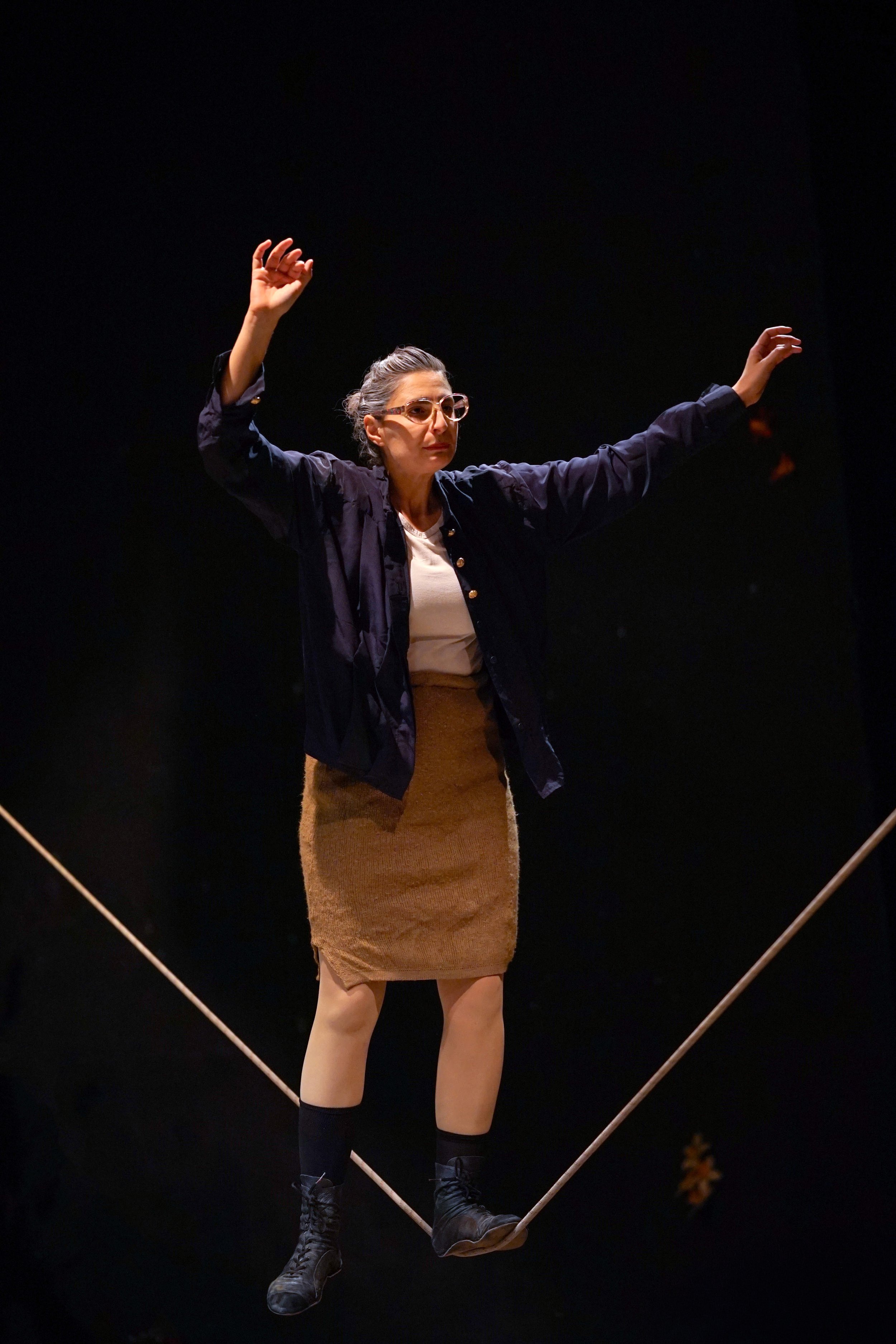 Donna in scena mentre cammina sull'attrezzo di un giro di equilibrismo, vestita con gonna marrone, maglietta bianca e giacca blu, con occhiali e capelli raccolti.