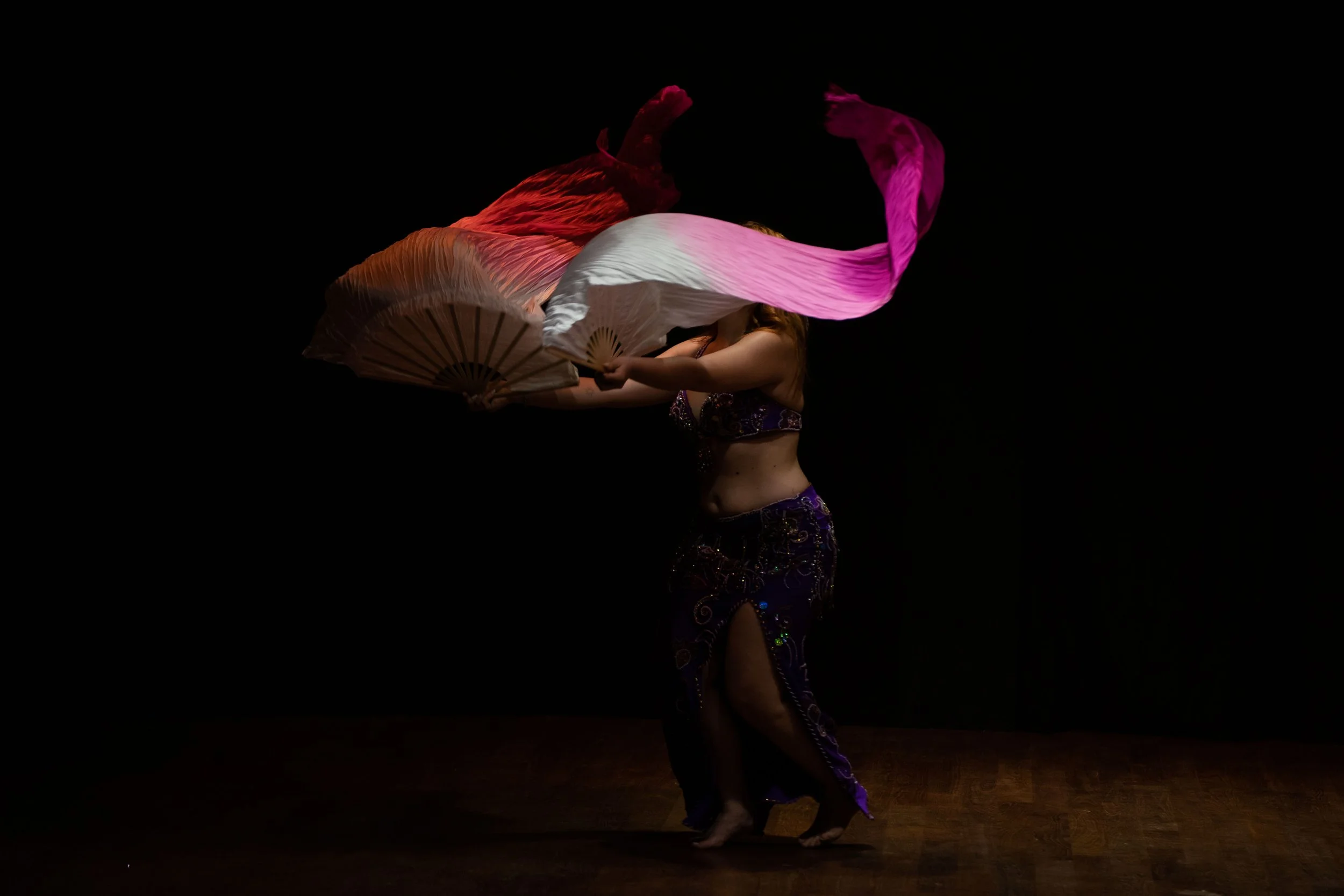 Danzatrice con copricapi e abiti colorati, che fa una performance con ventagli di stoffa variopinti in uno sfondo nero.