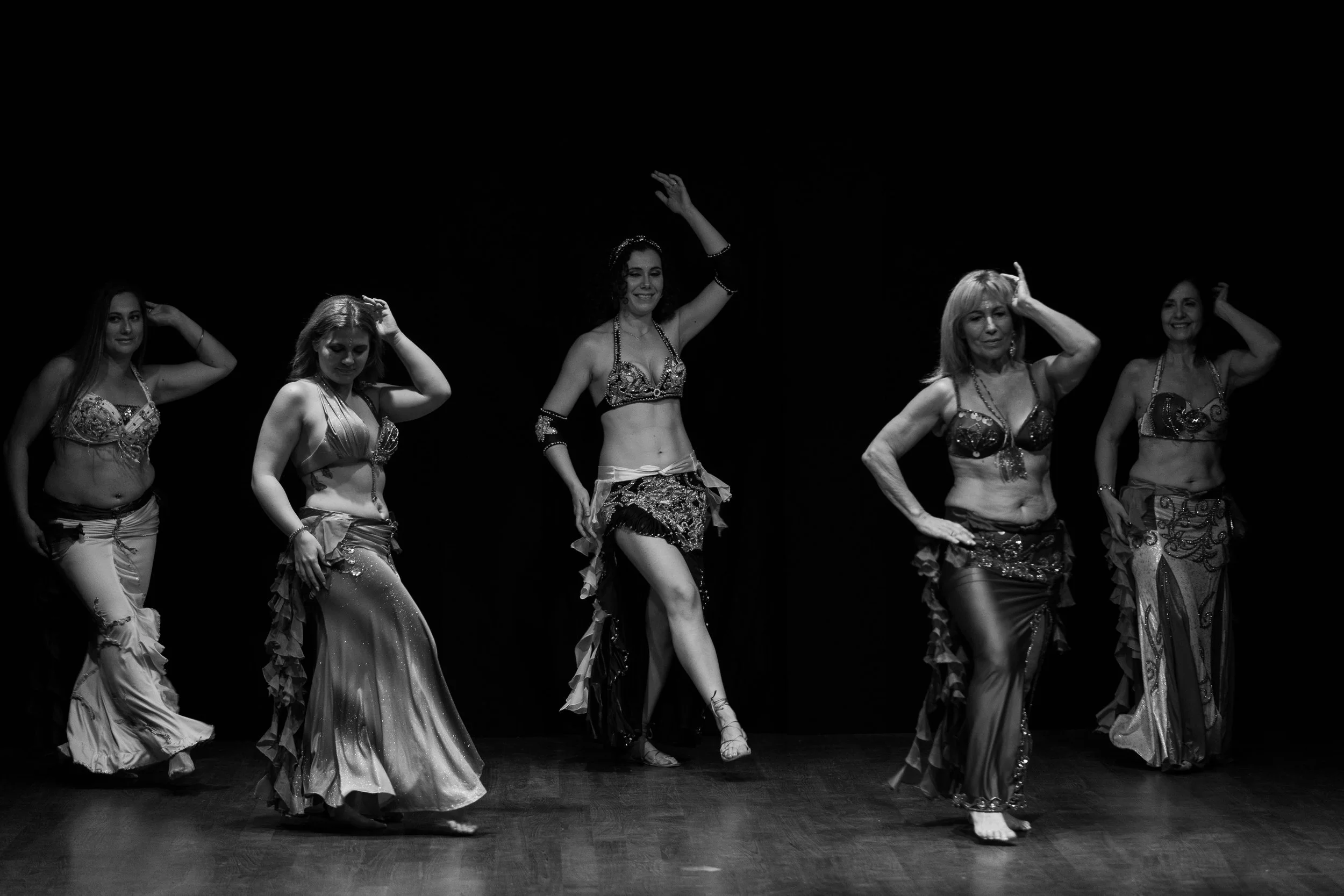 Cinque donne che ballano la danza del ventre su un palcoscenico, indossando costumi elaborati, in una performance in bianco e nero.