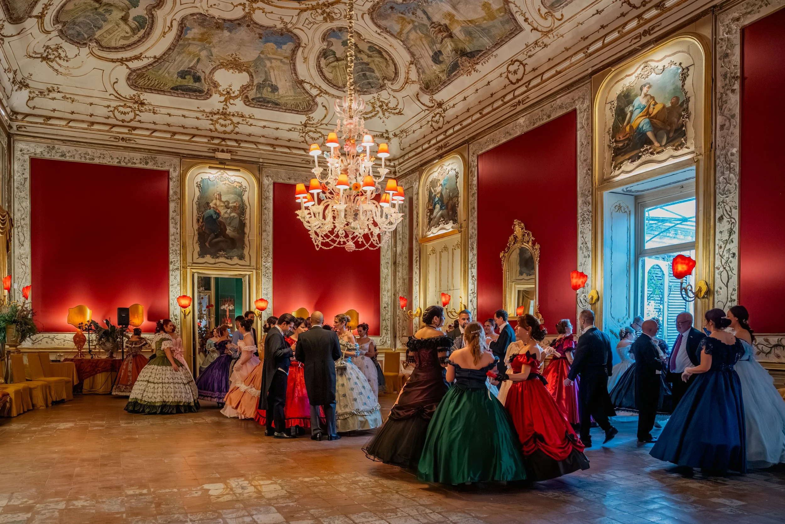 Sala elegante con soffitto decorato e grandi dipinti alle pareti, lampadario chandelier e numerosi ospiti in abiti formali e storici per un ballo o festa elegante.