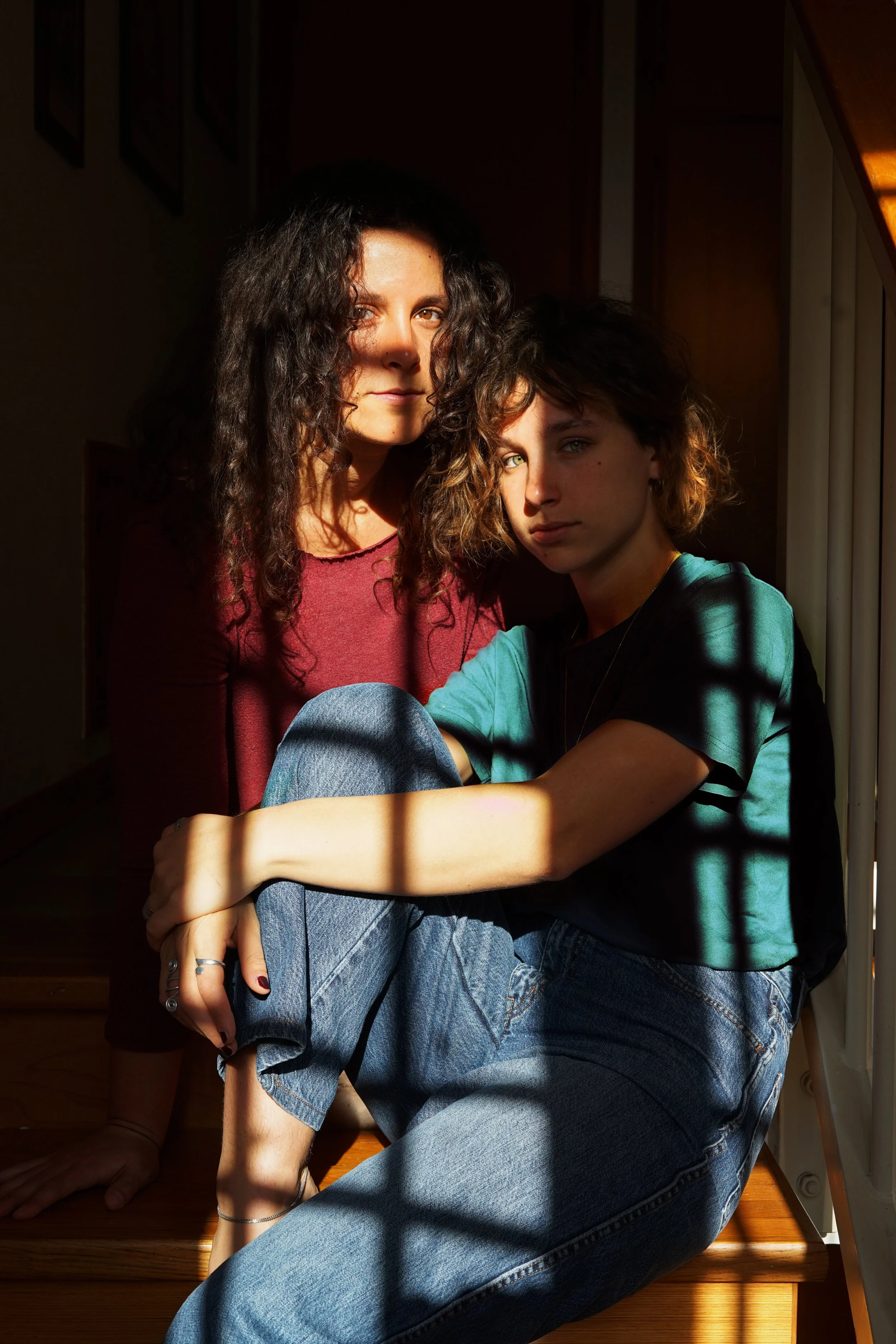 Due ragazze sedute vicino a una finestra, con raggi di luce che creano ombre sul loro volto e sul pavimento in legno.