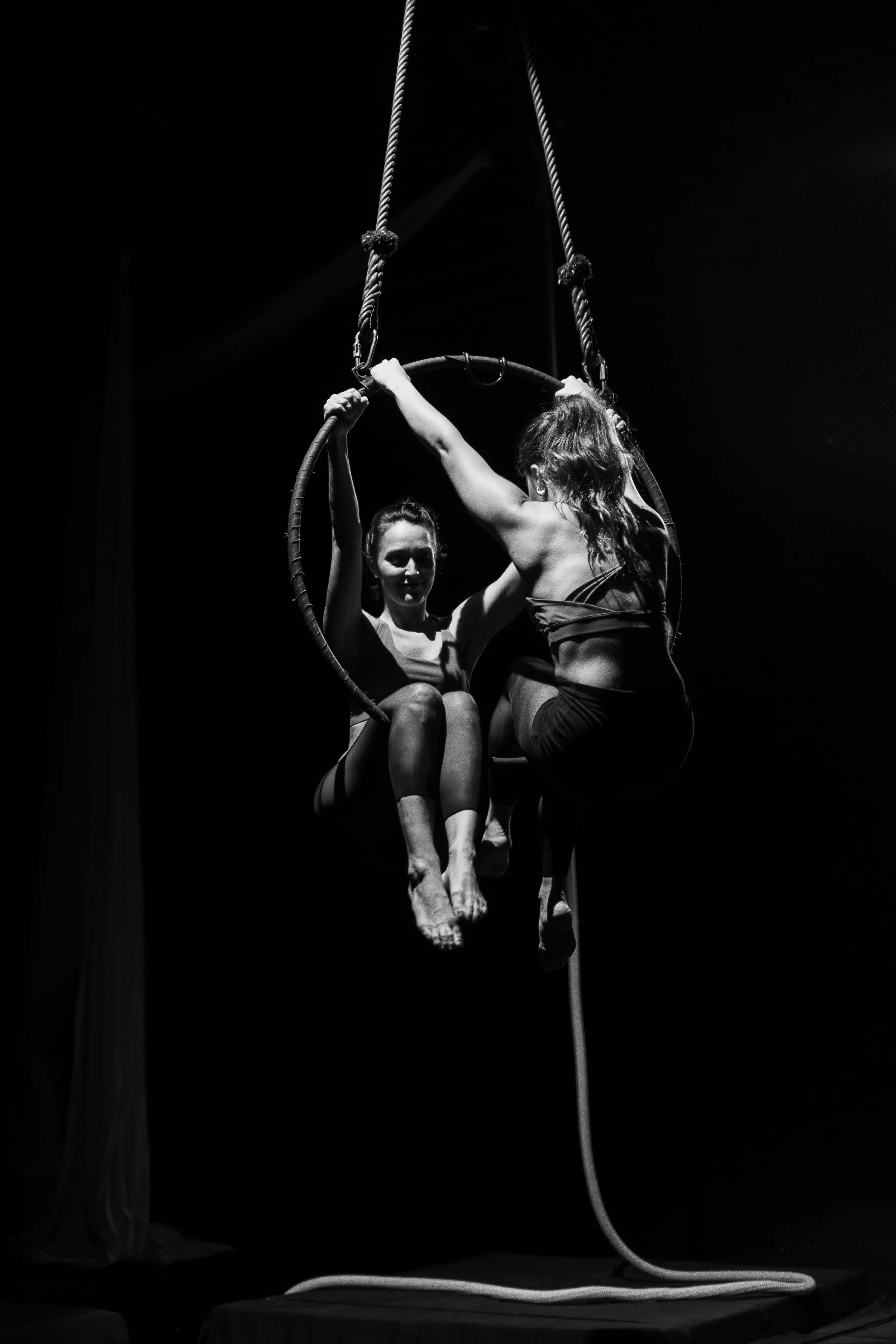Due donne praticano acrobazia su un cerchio sospeso con funi in un ambiente teatrale, illuminato in modo drammatico.